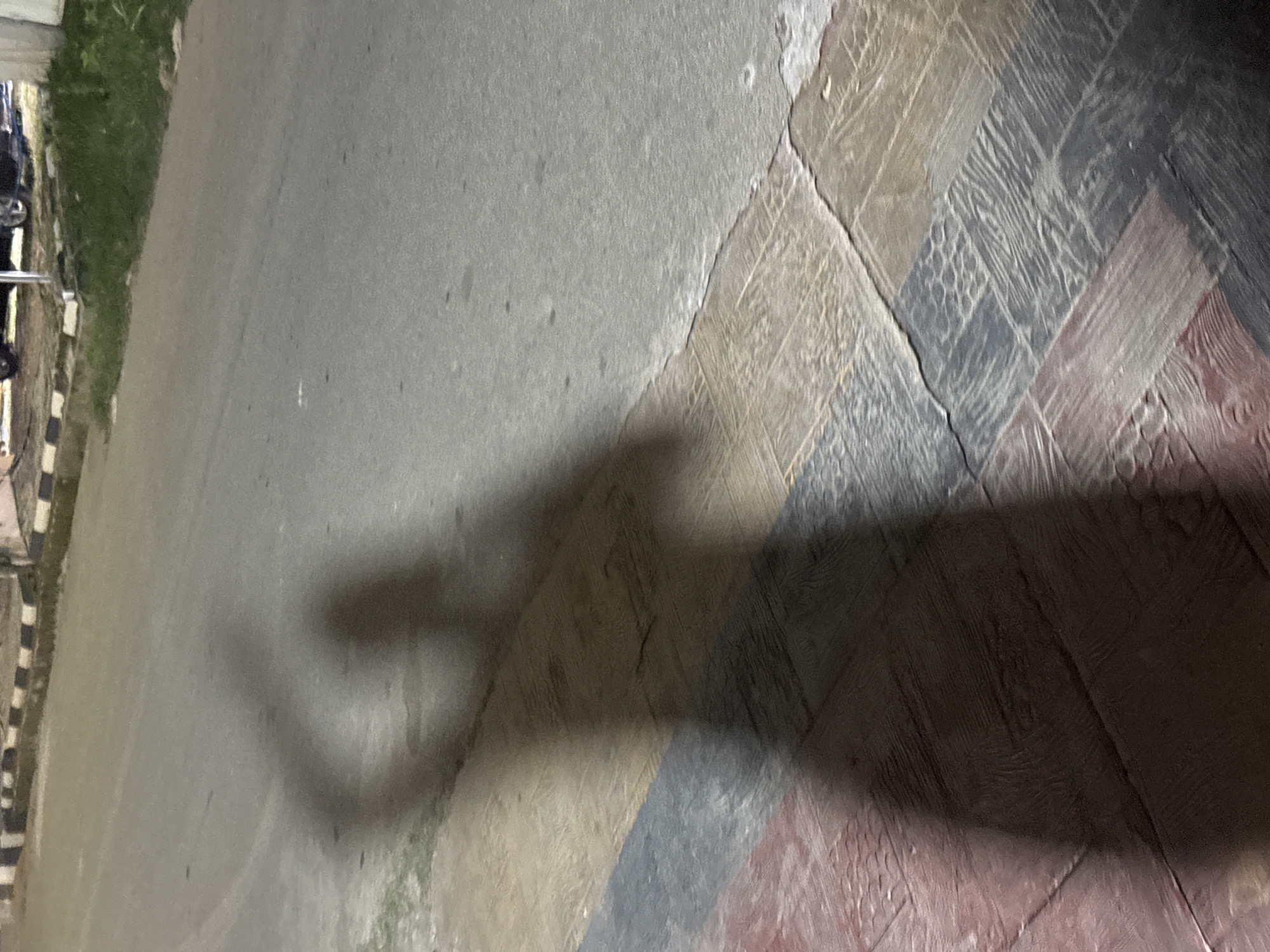 A long shadow of a person is cast on a paved surface with red, blue, and tan colored tiles. The shadow appears to be in motion, with one arm raised. The surface appears to be a sidewalk next to a paved road. In the background, there is a grassy area and what appears to be a curb with black and white stripes. There are also hints of vehicles in the background. The lighting suggests it is either dusk or dawn, or streetlights are on. The setting is likely an outdoor urban environment in Abuja, Nigeria.