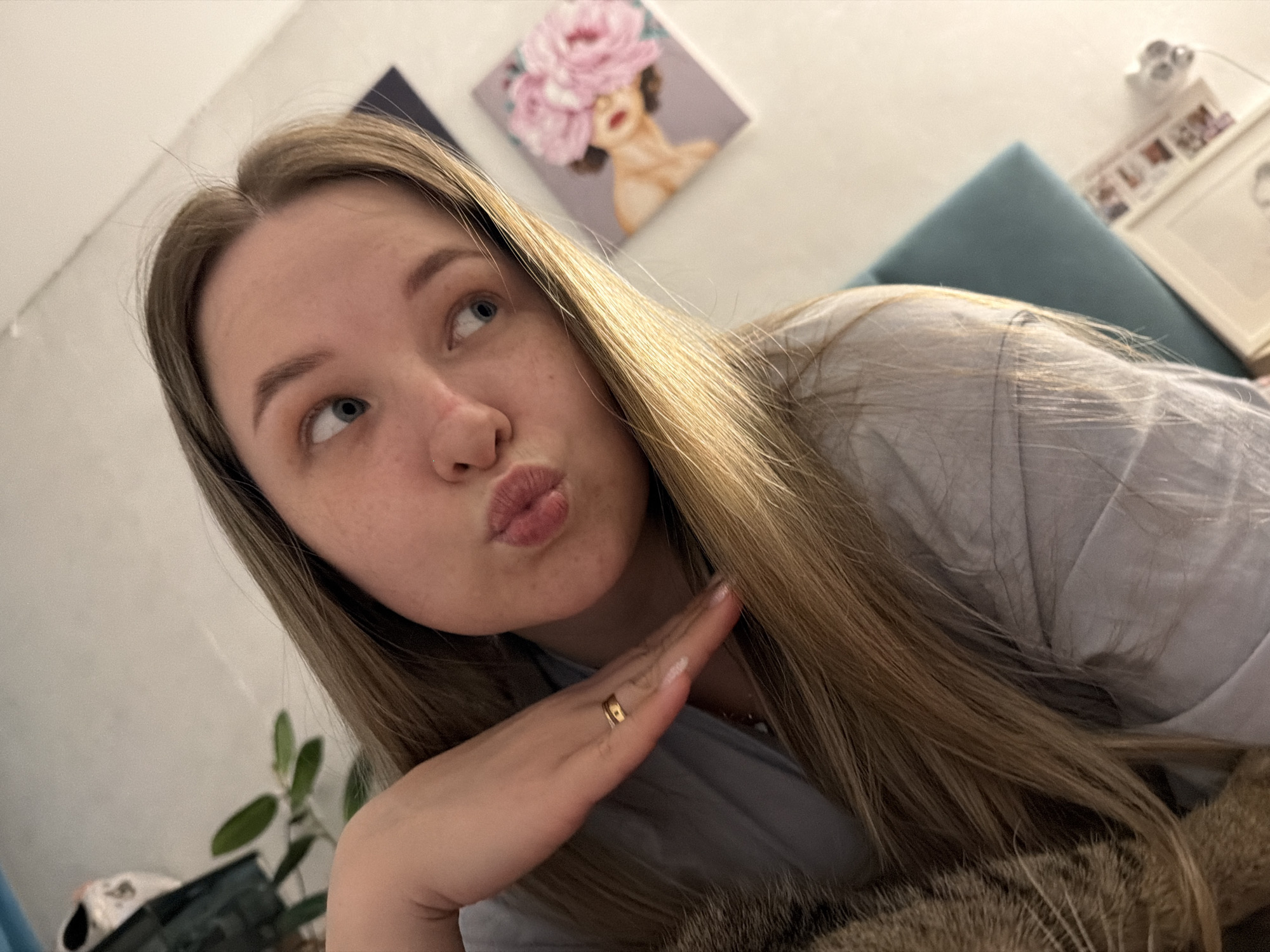 A young woman with long, blonde hair and fair skin poses indoors. She is looking up and to the left with a pouting expression. Her right hand is raised with her palm facing the camera, and a gold ring is visible on her ring finger. She is wearing a light grey shirt.
Behind her, on the wall, are two framed pictures. The first shows a stylized portrait of a woman with flowers in her hair. To the right, there is a second frame containing photographs. A teal-colored cushion is also visible behind her. A plant is partially visible in the lower left corner. A cat is partially visible, at the bottom right.
The location is the city of Chaikovsky, Russia.