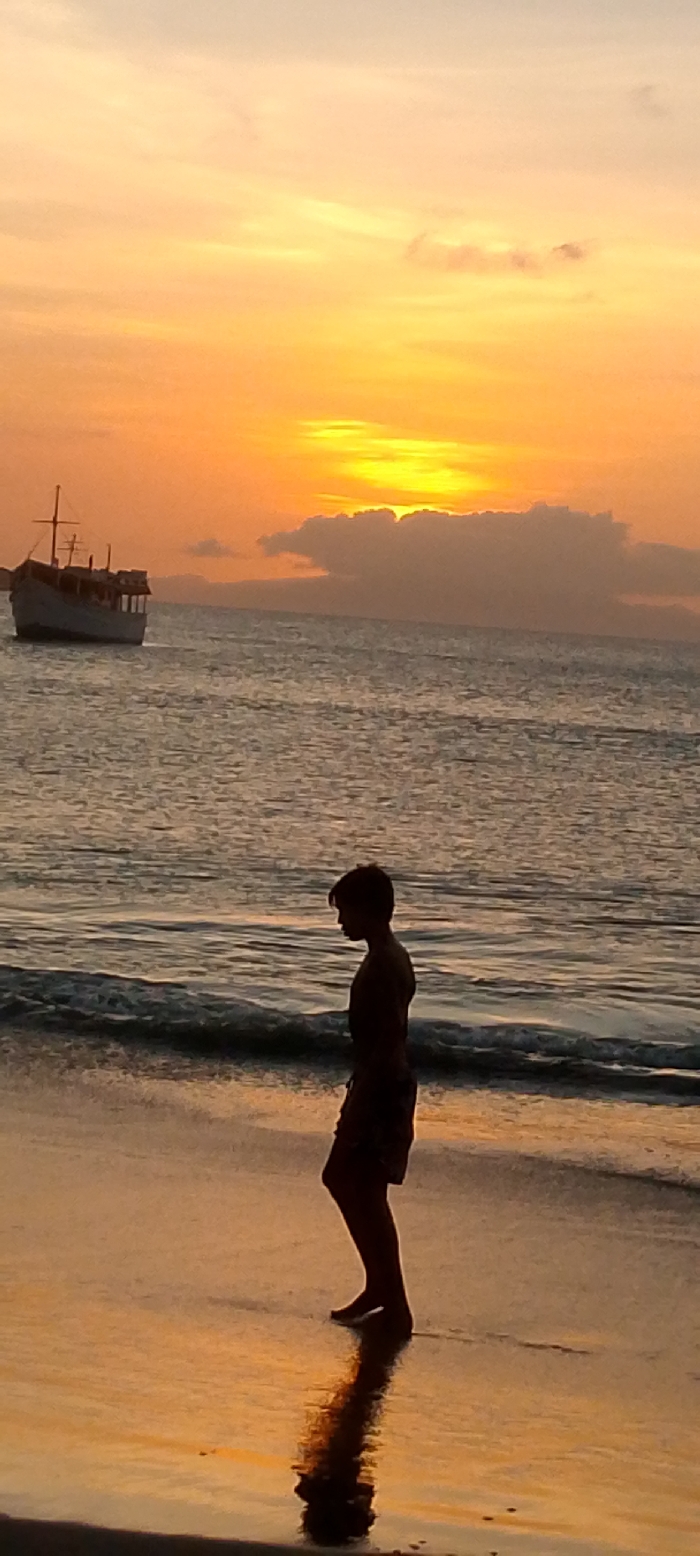 A silhouette of a person stands on a beach during sunset. The person is facing left, with their reflection visible in the wet sand. The sky is a gradient of orange and yellow, with the sun partially obscured by a cloud formation. A boat is anchored in the water, off to the left. The ocean waves are gently lapping at the shore. The scene evokes a sense of tranquility and solitude.