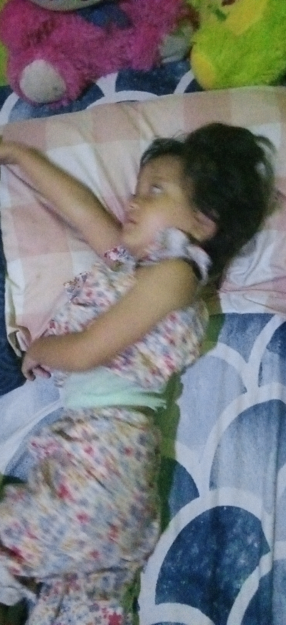 The image captures a young child lying on a bed, appearing to be asleep or resting. The child is positioned on their back, with their head turned slightly to the left, eyes closed or mostly closed, and one arm extended upwards over a pillow. They are wearing a light-colored, floral-patterned dress or sleepwear with a light green band around the waist. Their dark hair is spread around their head on the pillow.
The setting appears to be indoors, likely a bedroom. The child is on a bed covered with a blue and white patterned sheet. A light-colored pillow with subtle striped patterns (pink, white, and tan) supports the child's head. In the background, two stuffed animals are visible: a prominent pink teddy bear and a less distinct green stuffed animal, suggesting a child's sleeping area.
The overall impression is one of peaceful rest. Due to the indoor setting, the time of day and weather cannot be determined from the visual information alone. There are no specific visual cues within the image to indicate the provided location of Banjaran, Indonesia. The image is slightly blurry, particularly around the child's face and arms. There is no visible text in the media file.