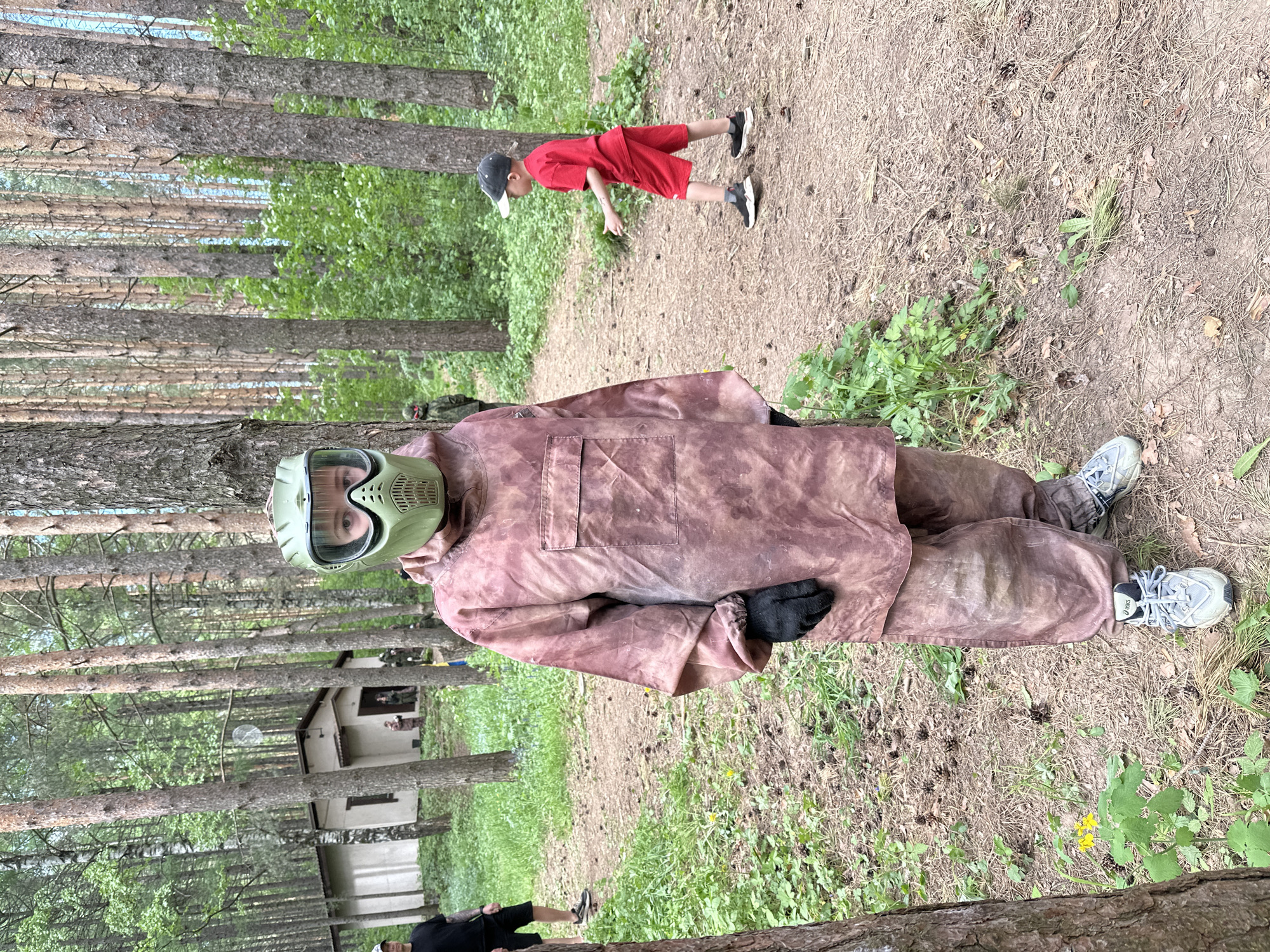 The image captures a scene in a wooded area, likely outdoors. In the foreground, a person wearing a camouflage jumpsuit and a protective face mask is lying on the ground. They have on gloves and sneakers. A young boy in a red shirt and shorts is seen standing further back on a path, and another person is partially visible at the bottom edge of the frame.
The background is filled with tall trees, suggesting a forest or park setting. A small building is visible in the distance. The ground is covered with dry leaves, pine needles, and patches of green vegetation.
While the image location is described as Votkinsk, Russia, there is no direct evidence within the image to confirm this location. This information might be from the context in which the image was provided rather than visible elements within the scene.