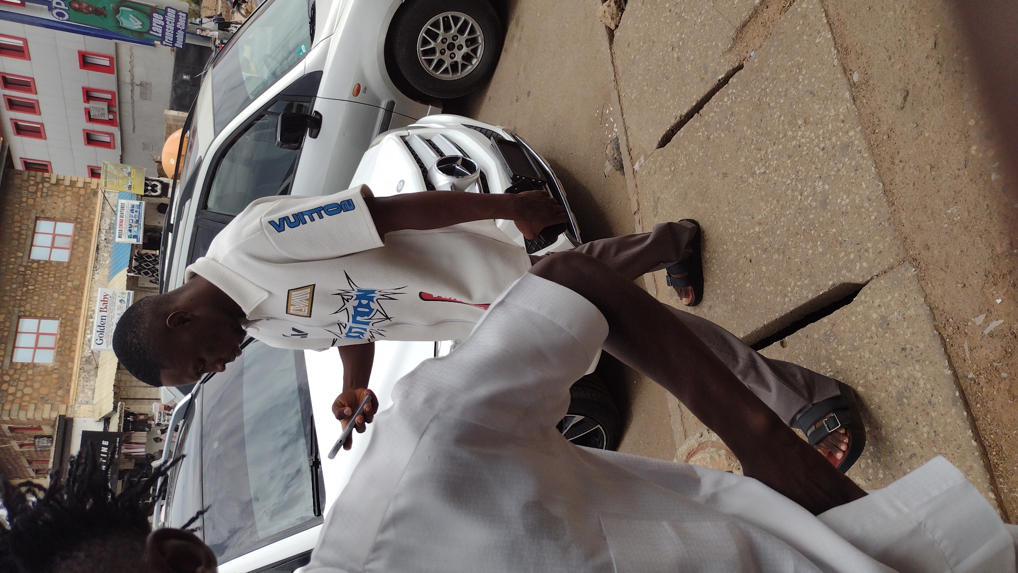 A street scene in Jos, Nigeria, shows two men standing beside a white car, identified as a Mercedes-Benz, with a modified front grill. One man, wearing a white collared shirt with "VUITTON" lettering on the sleeve and khaki trousers, is holding a smartphone. The pavement is cracked and uneven. Buildings are in the background, including a sign reading "Golden Baby." Additional commercial signage is visible.