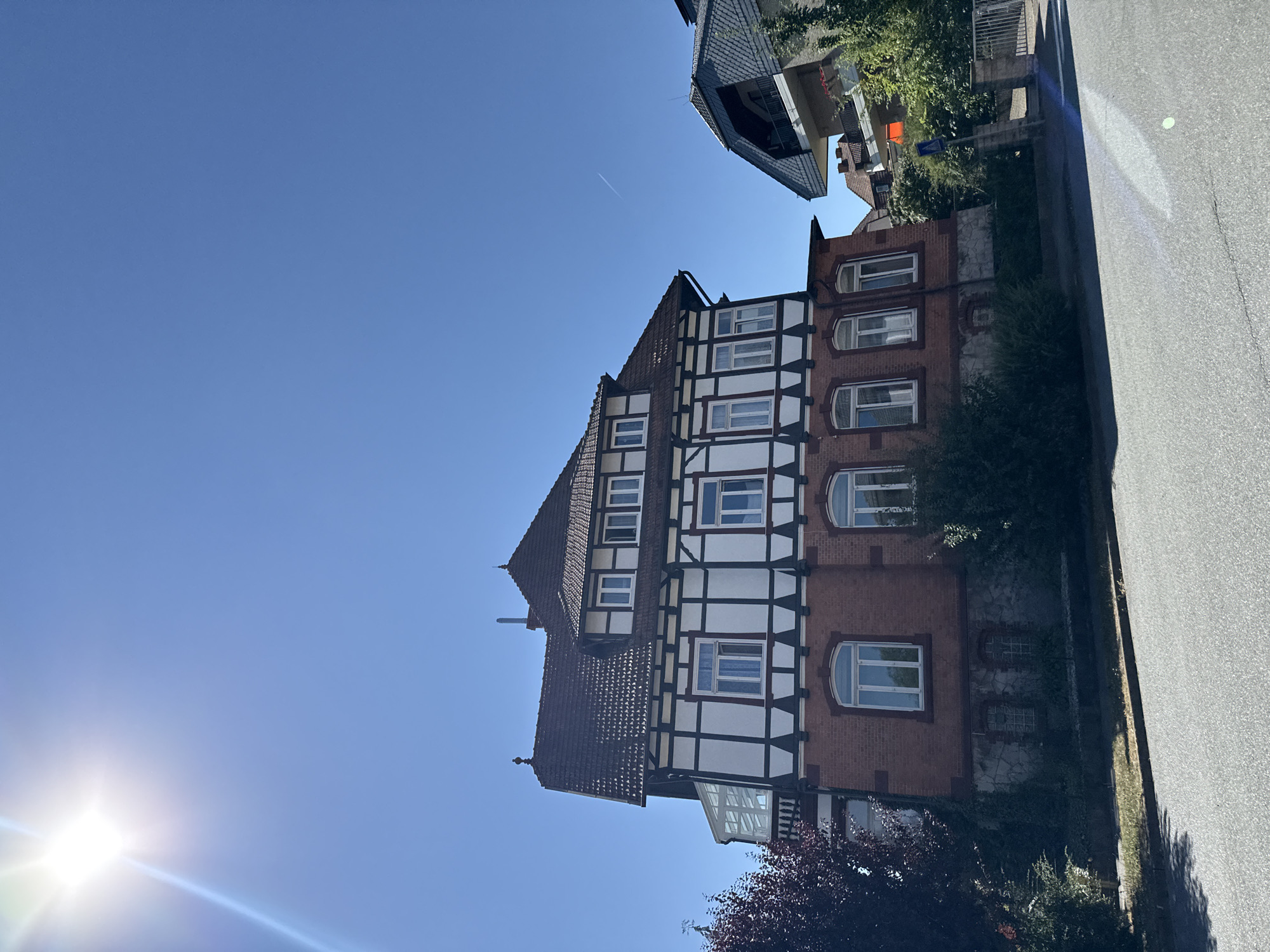 In Bad Sachsa, Germany, on a clear, sunny day, a traditional half-timbered house with a red brick lower story stands on a street. The house features white stucco panels bordered by dark timber framing, a dark tiled roof, and multiple windows. A sunroom addition extends from the left side of the house. Adjacent to it, another building with a steeply pitched, dark roof and balconies is visible. The street in front is paved, with a sidewalk and vegetation lining the property. The sun is shining brightly in the upper left corner, casting lens flare across the image. A contrail is visible in the blue sky above.