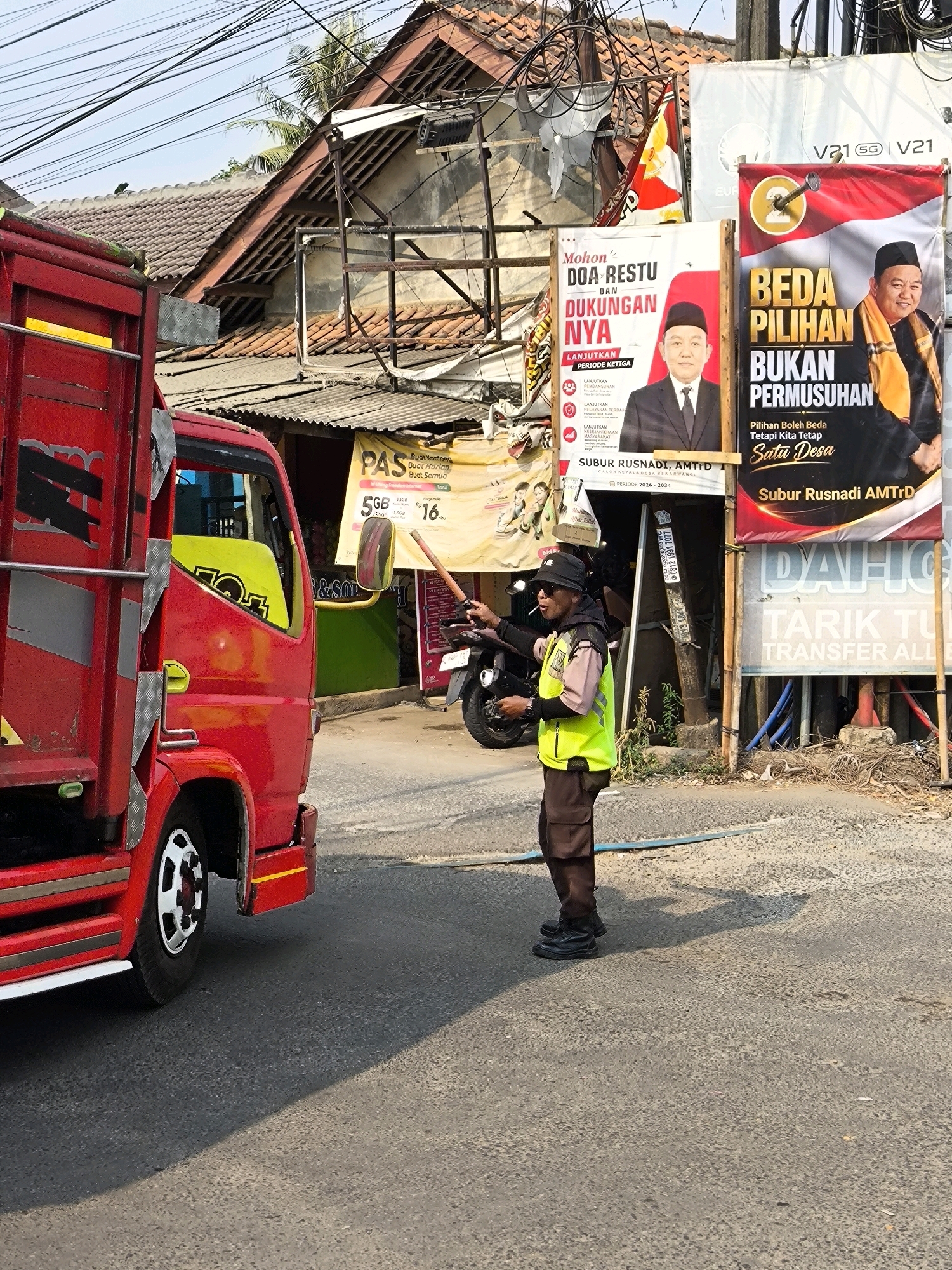 In the city of Lubangbuaya, Indonesia, on a sunlit day, a traffic officer in a neon yellow vest and dark pants directs traffic. He holds a wooden stick in his right hand, and appears to be smoking a cigarette. To his left is a red truck, its doors open. Behind the truck, a yellow sign displays the text "PAS" and "Buat Jajan" followed by "Buat Semua". Further in the background, a poster with a portrait of a man in a suit and a traditional cap, and the text "MOHON DOA RESTU DAN DUKUNGAN NYA LANJUTKAN PERIODE KETIGA" is visible. To the right of this, another poster features a man in traditional attire and the text "BEDA PILIHAN BUKAN PERMUSUHAN". The street is paved and dusty, with some debris on the side. Electrical wires are strung overhead, crisscrossing the sky.