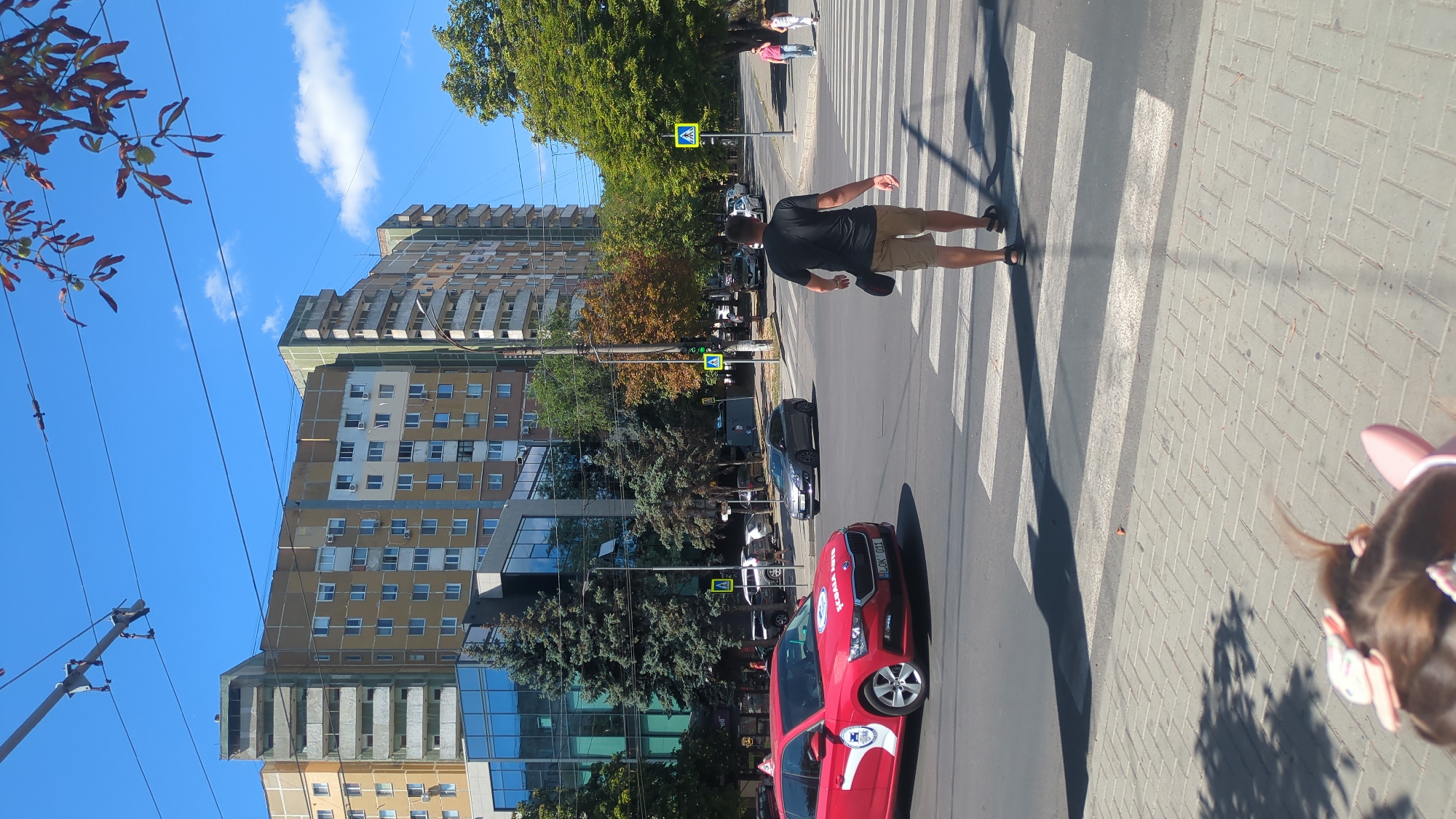 In this full-frame, eye-level, vertical shot of a sunny day in Chișinău, Moldova, a man is crossing a pedestrian zebra crossing. He is wearing a black t-shirt, khaki shorts, and black sandals, and carrying a dark bag over his right shoulder. His back is to the camera, and he is in motion.
To his left, a red Skoda Scala car, possibly a driving school vehicle with "SCOALA AUTO" written on its side, is stopped at the intersection. A silver sedan is also visible further down the road.
In the background, a modern glass-fronted building stands tall, flanked by mature evergreen trees. Behind this, a series of multi-story apartment buildings with balconies and numerous windows dominate the cityscape under a clear blue sky with scattered clouds. Overhead, power lines crisscross the sky, suggesting an urban environment. The street is paved, and pedestrian sidewalks are visible. The scene captures a typical moment in a city center, with traffic, pedestrians, and residential buildings under a bright sky.