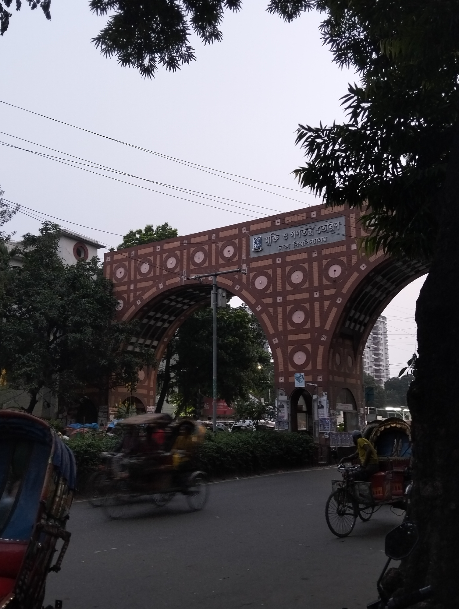 The image shows a street scene in Dhaka, Bangladesh, featuring the "Muktir Toron" (Gate of Freedom and Democracy) gate of Dhaka University. The gate is constructed of brick and has an arch design with circular and square patterns. The road in front of the gate shows motorized and non-motorized rickshaws in motion, indicating vehicular traffic. The scene is partially obscured by trees in the foreground. Additional buildings are visible in the background. Overhanging trees are visible at the top of the scene. The sky appears overcast.