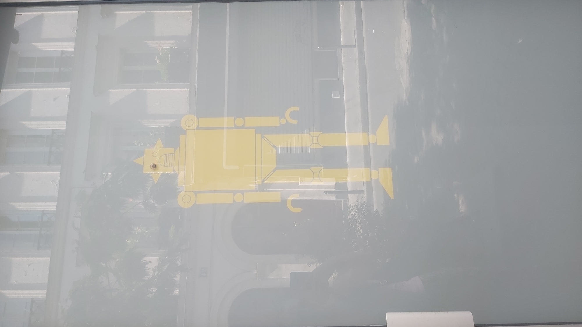 This image appears to be a photograph taken from inside a building, looking out through a window, possibly on a sunny day in Paris, France. The primary subject in the foreground is a large, yellow, graphic sticker of a robot adhered to the window. The robot sticker is stylized, with a square body, circular joints for the arms and legs, and a somewhat triangular head with two round eyes. The sticker is transparent enough that the background is visible through it.
The background shows a reflection and the actual scene outside. The reflection includes trees, buildings, and the sky, indicating a daylight setting. The actual scene outside the window reveals an urban environment with a building facade, featuring arched doorways or windows at street level and multiple stories with windows above. There are also trees and some greenery visible, suggesting a public space or a courtyard. The overall impression is of a bright, possibly slightly hazy or overexposed environment. The image is taken from a slight upward angle.
