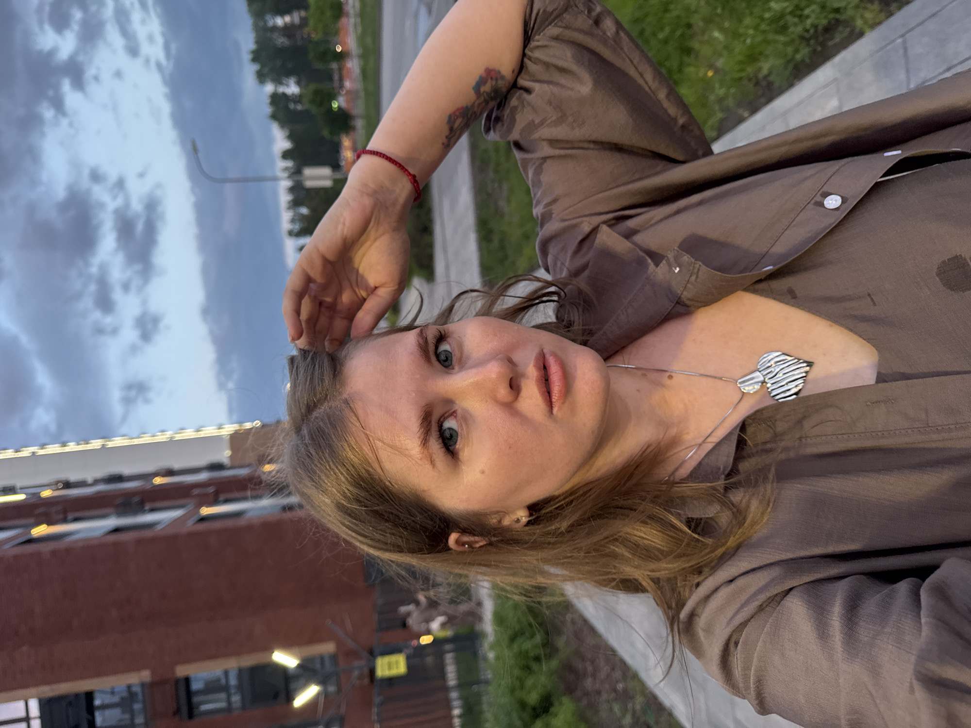 A woman takes a selfie outdoors in Perm, Russia. She faces the camera with a tilted head. She wears a brown button-down shirt, a silver heart-shaped necklace, and a red bracelet on her left wrist. A tattoo is visible on her left upper arm. Background features include a brick building with lit windows and exterior lighting, a sidewalk, green grass and foliage, and a street with a street light and other lit buildings in the distance. The sky is cloudy. A low metal fence is visible near the bottom of the frame.