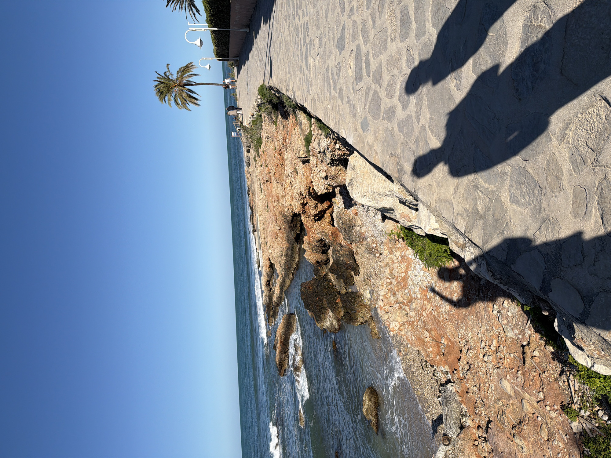 The image captures a coastal scene in Dénia, Spain. A stone pathway runs along the top right of the frame, casting shadows from people onto the path. Below, a rocky cliff descends to the sea, where waves crash against the rocks. A palm tree stands in the distance against a clear blue sky.