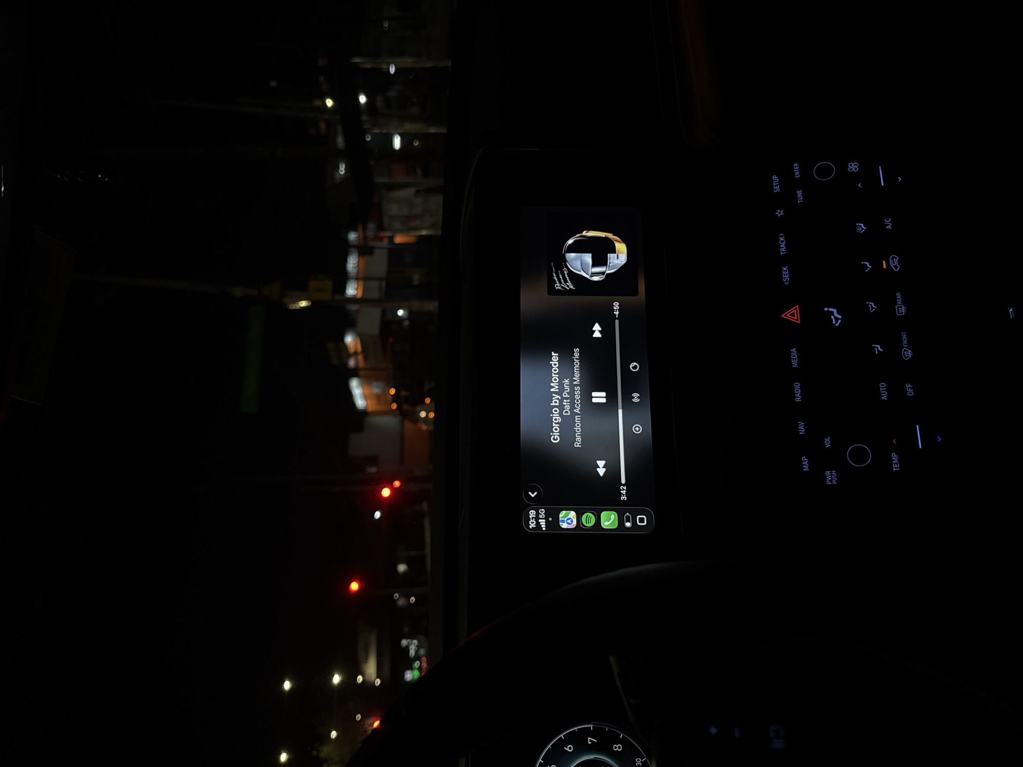 This media file captures the interior of a car at night, likely in Puebla, Mexico, from a driver's or front passenger's perspective, looking towards the dashboard and infotainment screen. The primary focus is the illuminated central infotainment display, which shows music currently playing.
The song displayed is "Giorgio by Moroder" by Daft Punk, from the album "Random Access Memories." The track is 3 minutes and 42 seconds into a 4 minute and 50-second duration. A small section of a connected smartphone interface is visible on the left side of the screen, indicating the current time as 10:19 and a "5G" cellular connection. Icons for Google Maps, Spotify, and another green app are present.
Below and to the right of the screen are various climate and media control buttons, also illuminated. These include labels such as "MAP," "NAV," "RADIO," "MEDIA," "TRACK," "SEEK," "SETUP," "TUNE," "ENTER," "VOL" (with a rotary knob), "PWR PUSH," "TEMP" (with an up arrow), "AUTO," "OFF," "FRONT," "REAR," "A/C," and an emergency hazard light button. Airflow direction indicators and a fan speed icon are also visible.
Through the windshield, the dark outside environment is visible, with numerous blurred, bright lights indicating streetlights, traffic signals (some red), and distant buildings, creating a bokeh effect. A portion of the steering wheel, showing numbers like "6," "7," "8," and "30" (likely part of a tachometer or speedometer) is visible at the bottom of the frame. The overall scene suggests a car either parked or moving slowly at night in an urban setting.