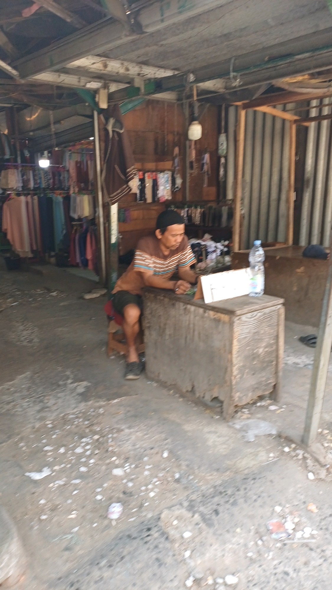A man wearing a black cap and a striped shirt sits at a wooden counter in an open-air market stall in Citeureup, Indonesia. He is leaning forward, looking at a white notepad or ledger on the counter. A bottle of water is placed on the counter to the right of the notepad. In the background, clothing is displayed on racks, suggesting a retail environment. The stall appears to be constructed with wood and corrugated metal. The ground is a concrete surface littered with small debris.