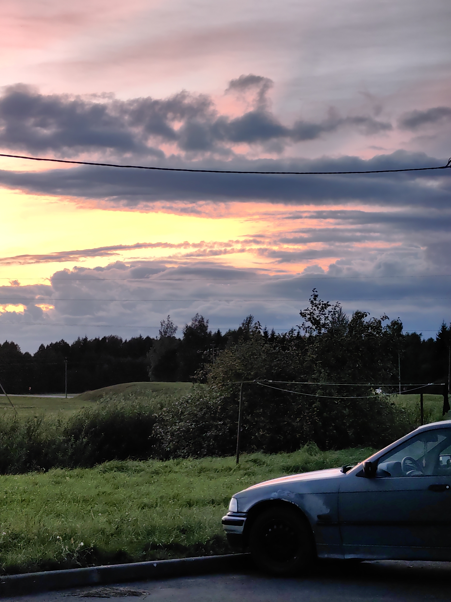 The image captures a twilight scene with a dramatic sky featuring pink and yellow clouds interspersed with dark grey formations. A dark wire runs horizontally across the frame, dividing the sky. Below the sky is a dark silhouette of a dense forest. In the foreground, a grassy embankment is visible, with a portion of a dark colored car, likely a sedan, parked on the right side of the frame. The car appears to be older and is facing left. The overall environment suggests a rural or semi-rural setting, possibly on the outskirts of the city of Myadzyel, Belarus, given the presence of trees and open fields under a prominent sky. No people are visible in the image.