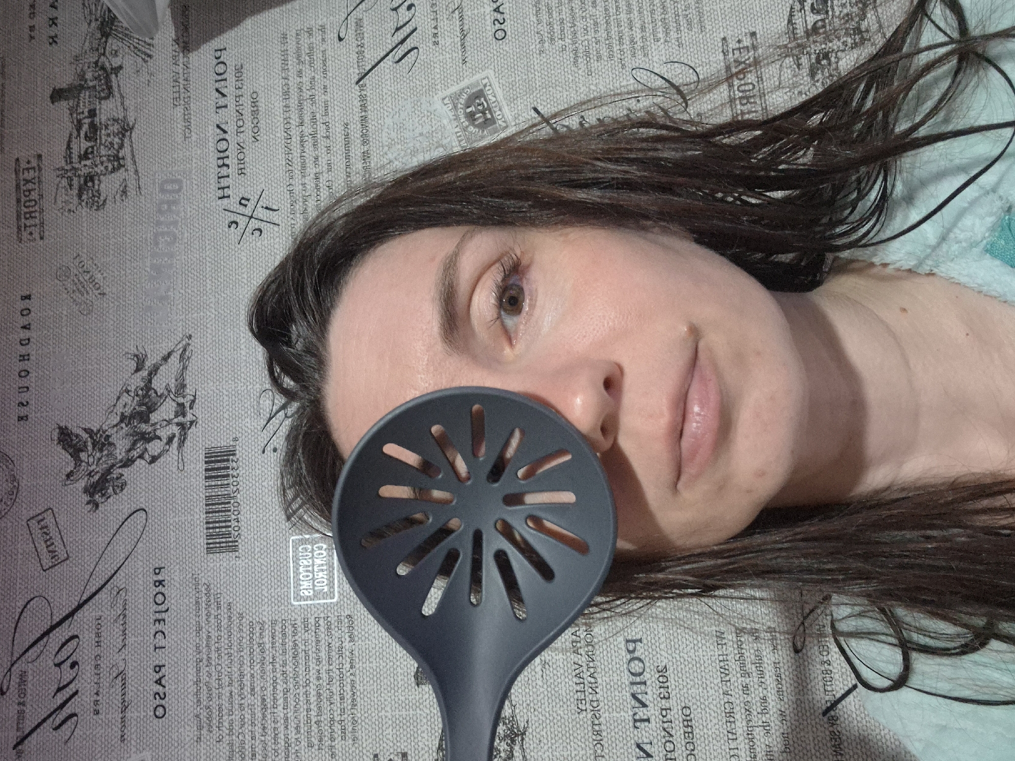 The image shows a person lying down with a black slotted spoon covering part of their face. The person has brown hair and fair skin. The background is a gray surface with various texts and graphics printed on it, reminiscent of a newspaper or printed fabric.