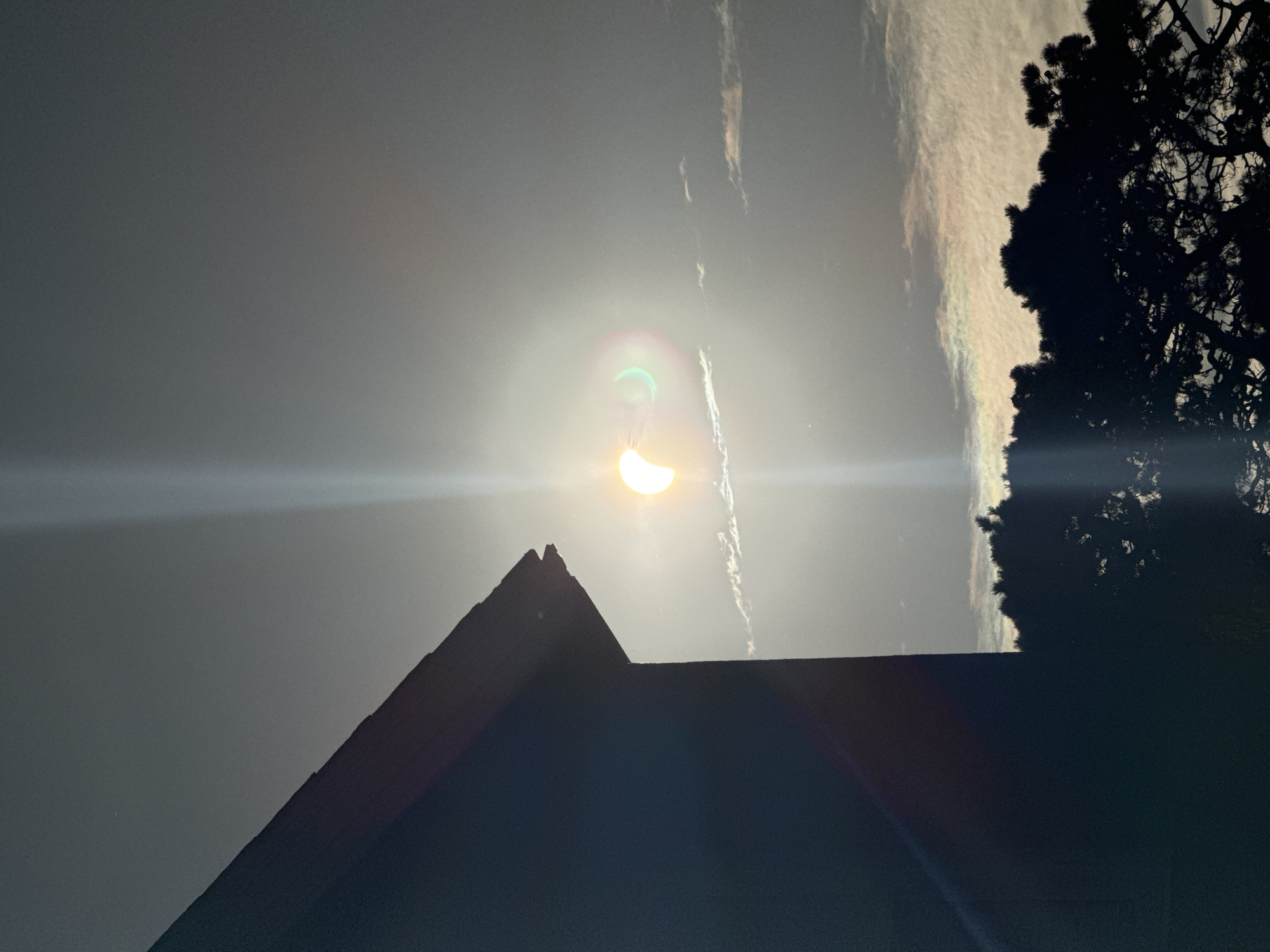 The image depicts a partial solar eclipse viewed from an urban environment, likely Waterford City, Ireland. In the foreground, the dark silhouette of a building's roofline cuts across the left side of the frame. The sky is filled with a hazy, overcast light, with the sun, partially obscured by the moon, shining brightly near the center. A prominent lens flare streaks vertically from the sun, creating a bright white column. To the right of the sun, a green and pink iridescent halo is visible, likely a result of light scattering through atmospheric particles or moisture. Wispy clouds are scattered across the sky, with some catching the light and appearing iridescent as well. In the lower portion of the image, the dark silhouette of a dense evergreen tree is visible, its branches reaching upwards. The overall atmosphere is one of muted light and celestial observation.