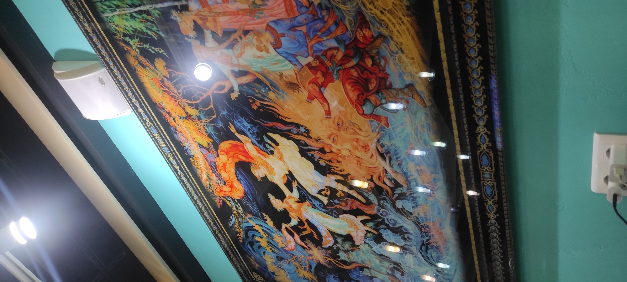 The image shows a large, ornate painting displayed on a turquoise wall in what appears to be an indoor setting. The painting depicts a vibrant, chaotic scene with figures in motion, possibly representing a mythological or historical event. The style is reminiscent of Persian miniature paintings, with intricate details, rich colors, and a black background. There are multiple figures in elaborate costumes, some appearing to fly or float amidst flames and swirling blue and white elements. A prominent, large, fiery-orange shape dominates the upper left of the painting, possibly a dragon or a stylized representation of fire or energy.
The painting is framed by a dark border with decorative gold and blue patterns. Above the painting, a white speaker is mounted on the wall, and a strip light illuminates the scene from the ceiling. The top of the frame is angled upwards, suggesting the painting is mounted high on the wall.
The lower portion of the image shows the turquoise wall extending downwards. At the very bottom, a white electrical outlet with two plugs is visible, with a black cable extending from it.
Given the information provided, this scene is not located in Moscow, Russia. The image context and visual cues do not suggest a Moscow location.