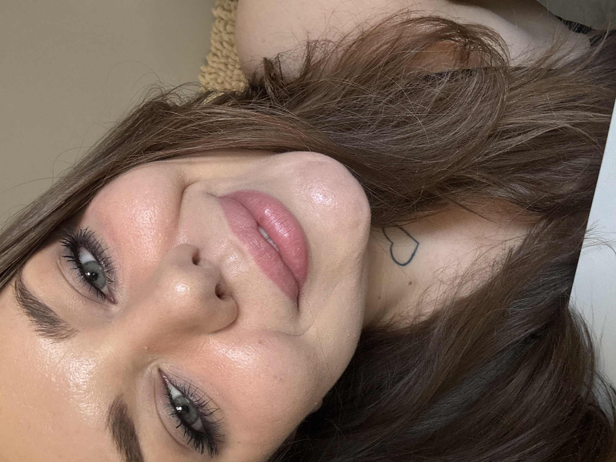 The image is a close-up selfie of a young woman's face. She has brown hair, blue eyes, and is wearing makeup, including mascara and lipstick. A small, outlined heart tattoo is visible on her neck. The background appears to be indoors with a textured surface. The image is taken from a slightly upward angle, showing her chin and neck prominently. No specific location in Kremenchuk, Ukraine, can be identified from the image.