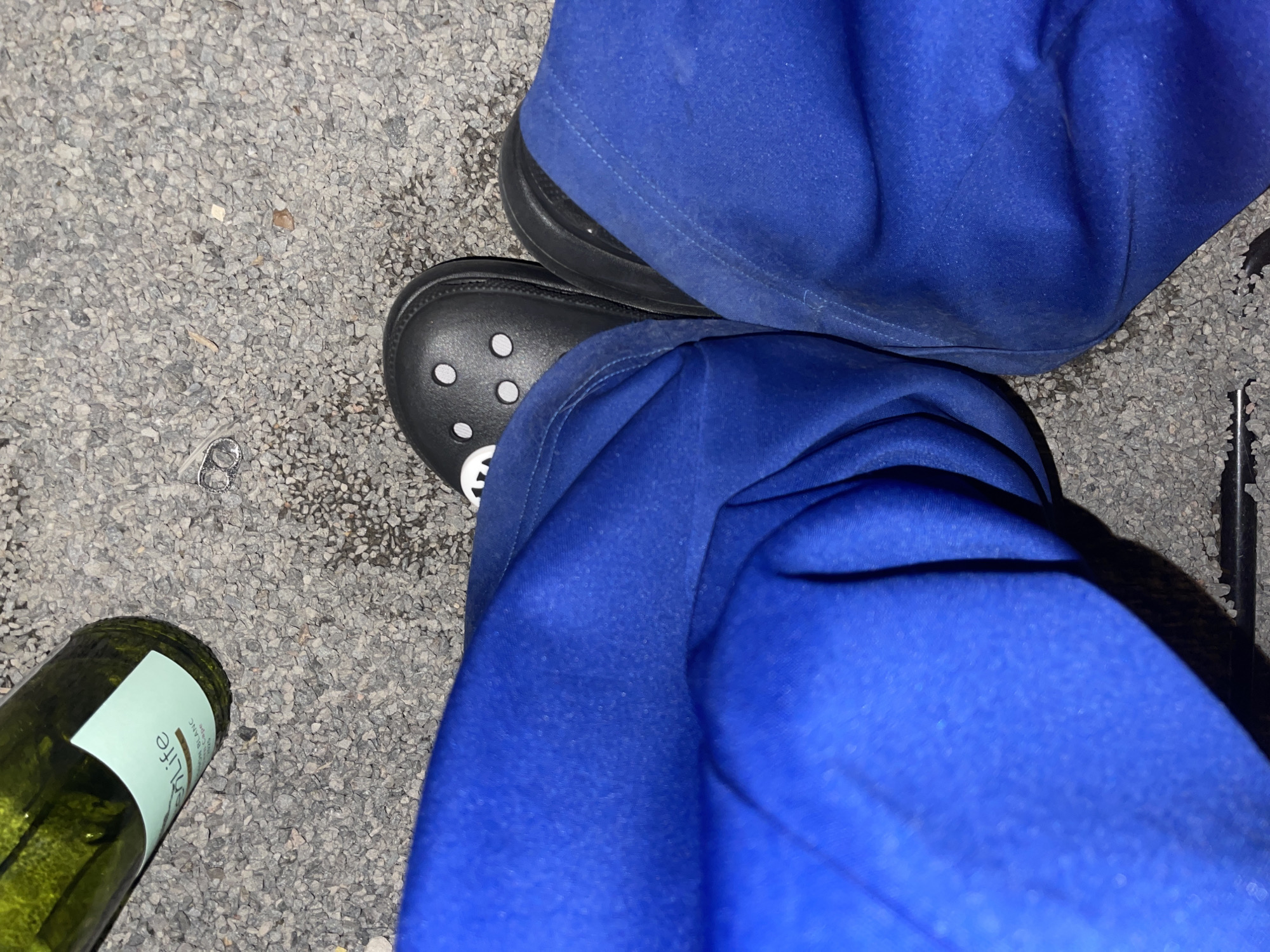 The image shows a close-up view of a person's legs wearing blue scrubs and black Crocs, standing on a gravel surface. A green wine bottle with a light blue label is visible to the left, resting on the gravel. The wine bottle label reads "life" and "MON BLANC". The scene is outdoors, on a surface that appears to be asphalt or a similar ground material covered in small stones. The overall lighting suggests it is either nighttime or a dimly lit environment. No specific location within Khimki, Russia, is identifiable from the image.