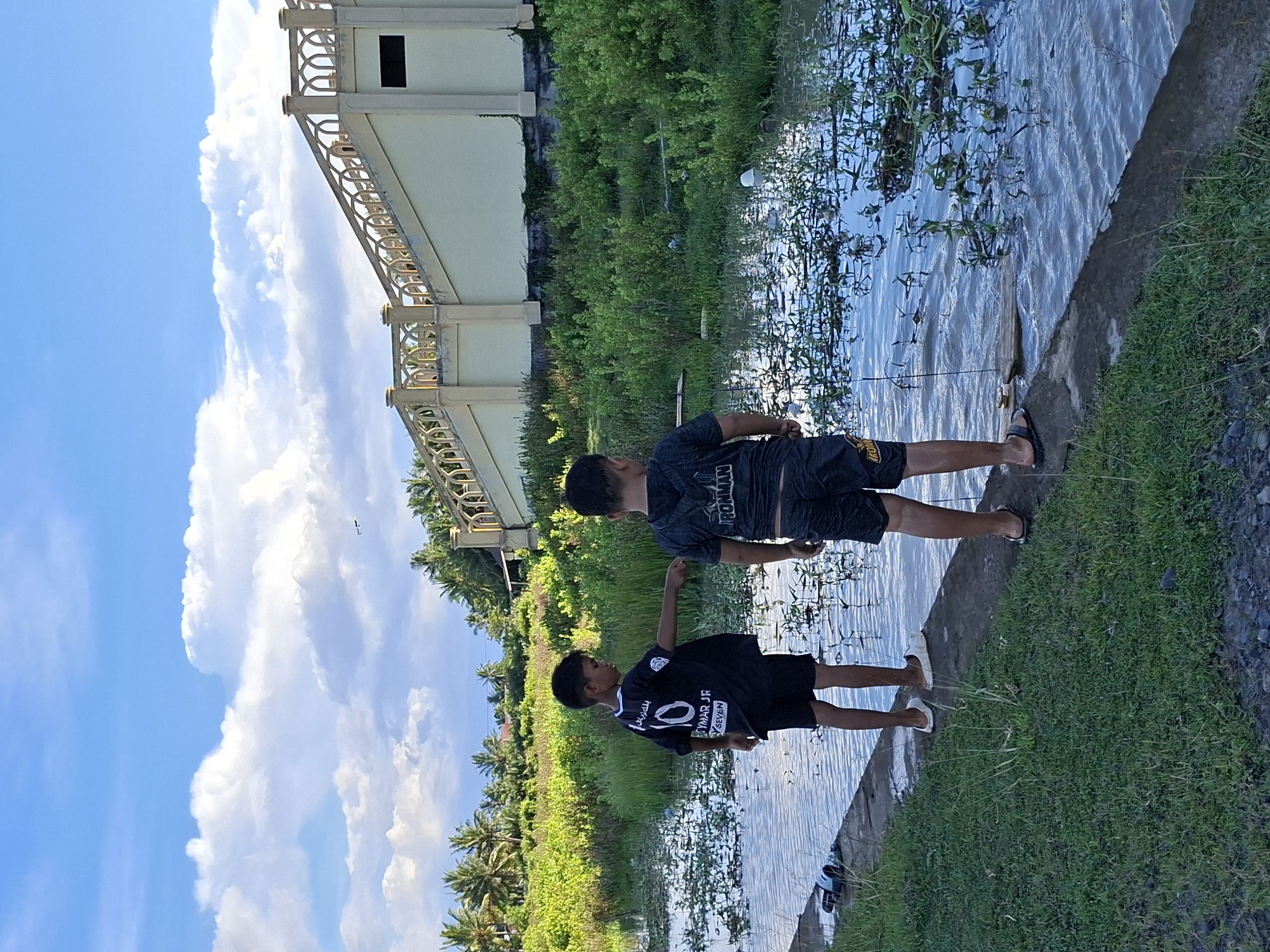 Two boys are fishing in a canal in Keudegeudong, Indonesia. The older boy is wearing an "Ironman" shirt and black shorts, while the younger boy is wearing a soccer jersey with "Neymar Jr." printed on it. They are standing on a concrete ledge, with water plants growing in the canal. Lush greenery and palm trees line the canal's edge. A two-story building stands nearby. The sky is blue with scattered clouds.