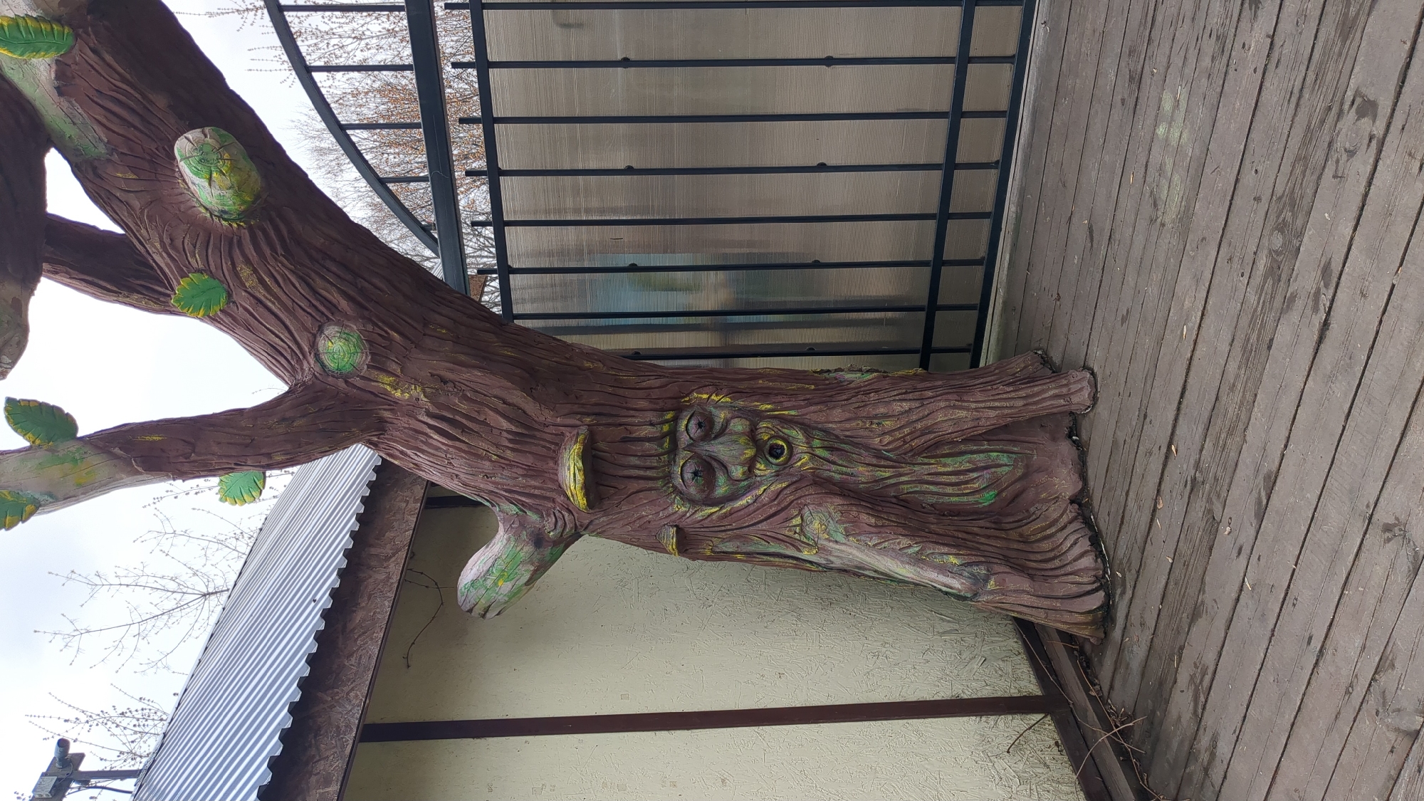 The image depicts a faux tree with a face carved into its trunk, located outdoors in what appears to be the city of Ulyanovsk, Russia. The artificial tree, painted in shades of brown with touches of green and yellow, is positioned next to a building with a corrugated metal awning and a white wall. Above the tree, there's a structure with a black metal frame and a translucent roof. The "tree" is standing on a wooden deck. The sky is visible in the background.
