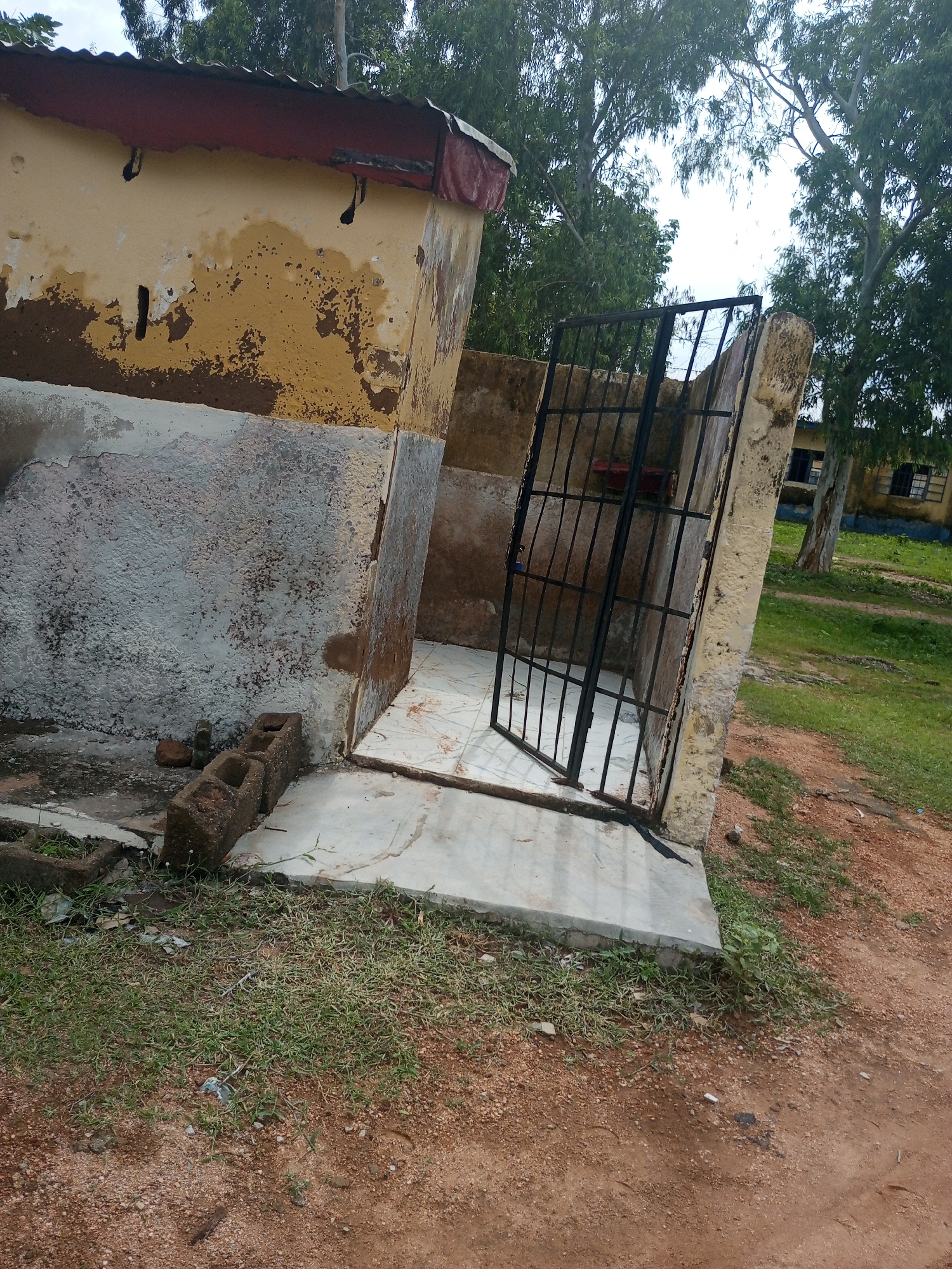 The image depicts an outdoor scene in Jalingo, Nigeria, featuring a dilapidated building with peeling yellow paint and a rusted corrugated metal roof. The building appears to be part of a larger structure, with a wall extending from it, also showing signs of decay. In front of the building, there is a small enclosed area with a metal gate. The ground is a mixture of concrete, dirt, and sparse grass. Several hollow concrete blocks are stacked near the building. In the background, tall trees with dense foliage are visible, and beyond them, a building with windows can be seen, suggesting the presence of a school or similar institution. The sky is overcast, indicating a cloudy day. There is no discernible activity or event taking place; the scene appears to be static and abandoned. The overall impression is one of neglect and disrepair.