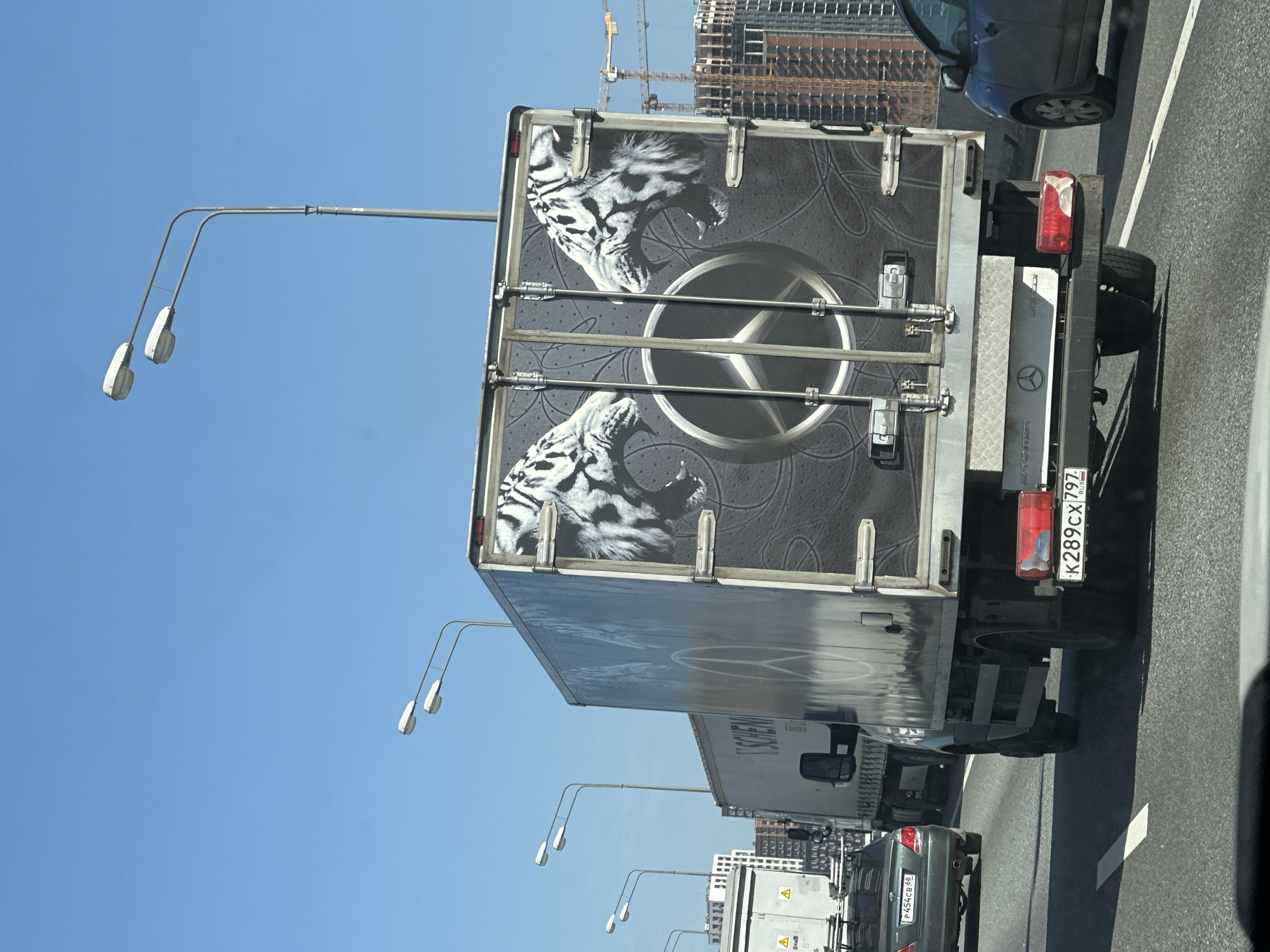 The image shows the back of a Mercedes-Benz Sprinter van with a striking design of two white tigers roaring. The van's license plate reads "K289CX797 RUS." The background reveals a city environment with cranes and buildings under construction, suggesting a large city skyline. The van is on a highway, likely in Moscow, Russia. Several cars are visible, as well as rows of street lights.