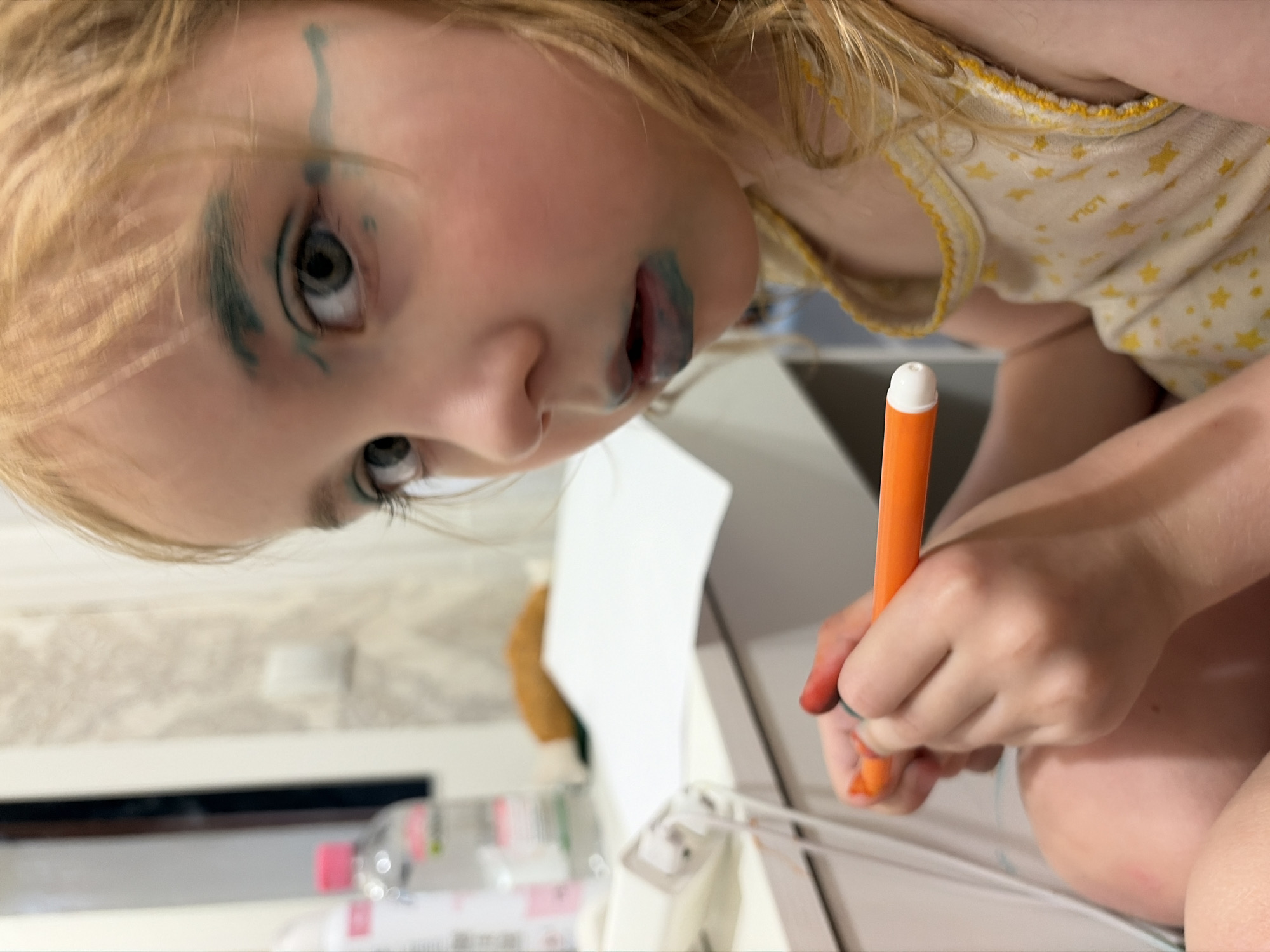 The image shows a close-up, portrait-style, sideways shot of a young child with blonde hair, who appears to be around 3-5 years old. The child's face is partially covered in blue marker, with marks on their eyelids, eyebrows, and lips. Their eyes are open and looking towards the camera. The child is wearing a light-colored, short-sleeved garment adorned with small yellow stars and a yellow trim, which has the word "LOLA" printed on it. In the child's right hand, they are holding an orange marker with a white tip, and their fingers are stained with red and blue ink. The child is positioned over a white surface, possibly a table or desk, where a sheet of white paper is visible. In the background, out of focus, there's a partially visible plastic bottle with a pink cap. The setting appears to be indoors, with a patterned wallpaper visible in the upper left background. The overall impression is of a child engaged in creative play, perhaps drawing or applying makeup, with a sense of childish mischief. The lighting suggests daytime, possibly indoors in a well-lit room.