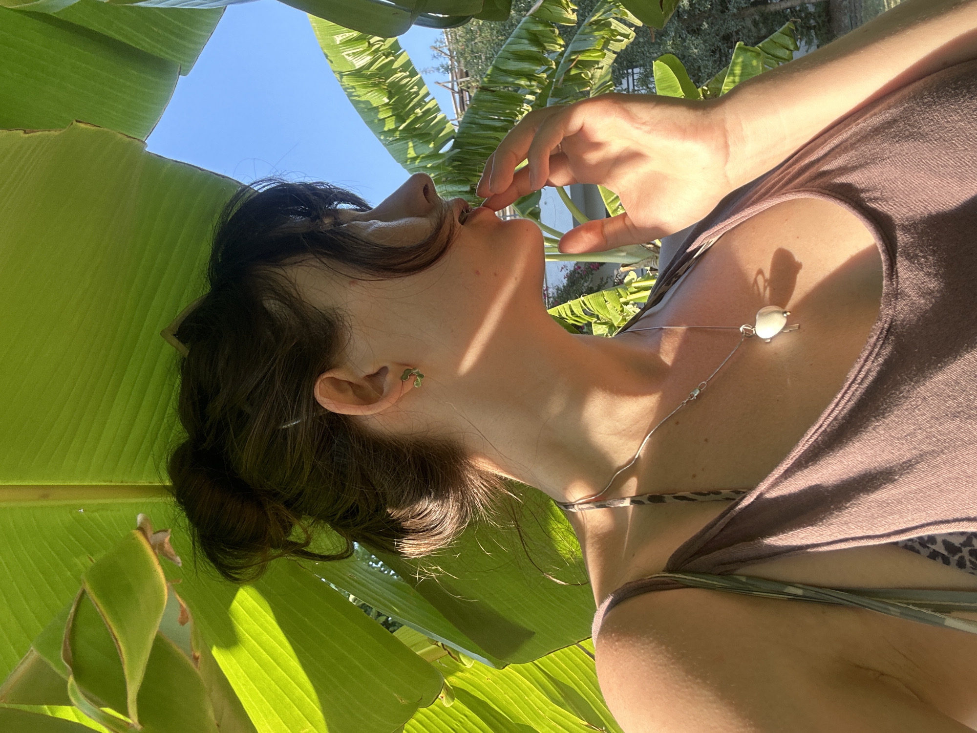 In Ortaca, Turkey, a young woman with dark hair pulled into a bun is seen from the chest up, partially obscured by large green banana leaves. She is looking to her right, with her right hand touching her lips. She is wearing a mauve tank top, a silver necklace with a heart-shaped pendant, and small green clover-shaped earrings. The background consists of a clear blue sky and some greenery. The lighting suggests it is daytime.