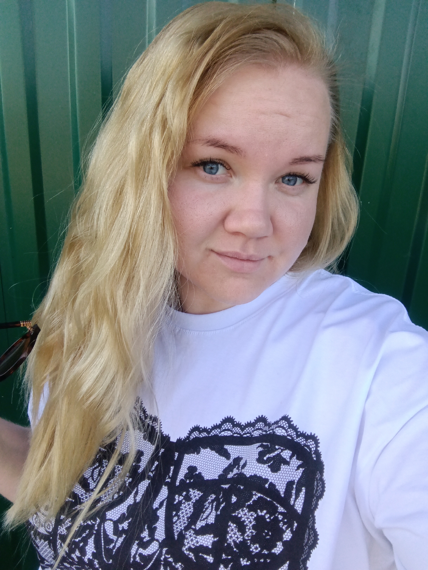 A blonde woman with blue eyes is centered in the frame, wearing a white t-shirt with a black lace pattern on the front. She is smiling slightly at the camera. Her hair is parted to the side and flows down her left shoulder. Part of a pair of sunglasses is visible to the left of her face. She is standing in front of a green corrugated metal wall. The image was taken in Фоки, Russia.