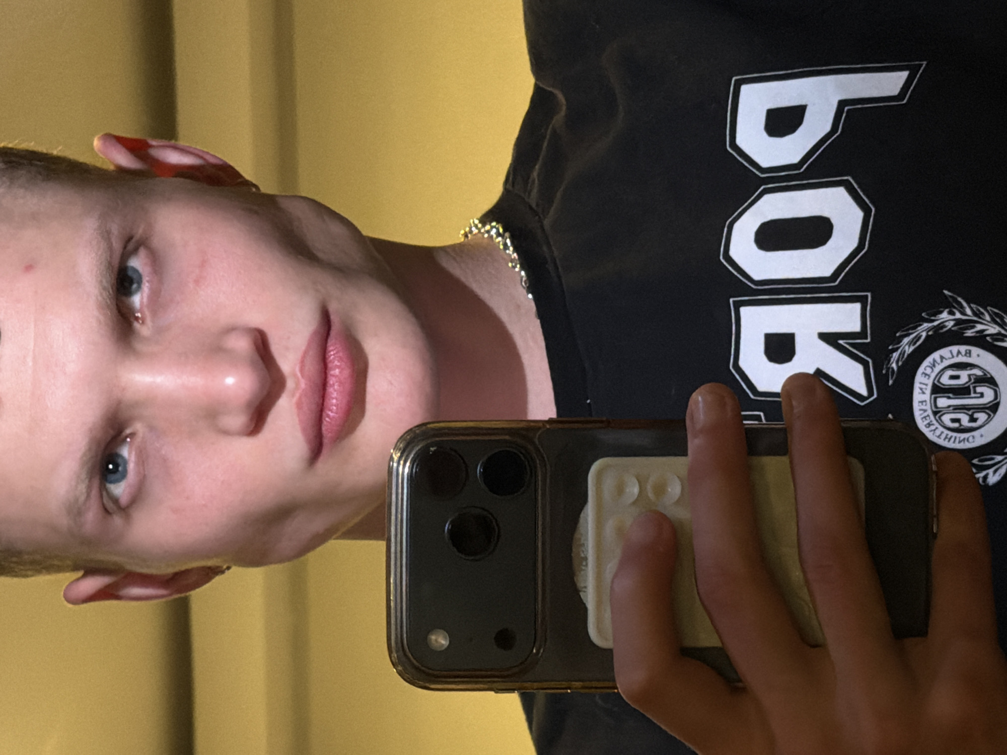 A light-skinned person with blue eyes is holding a smartphone to take a selfie. The phone has a dark-colored case and a white grip on the back. The person is wearing a black T-shirt with white lettering, and a thin gold chain is visible around their neck. The background appears to be a light yellow or beige wall. The image provides no information about the city of London.