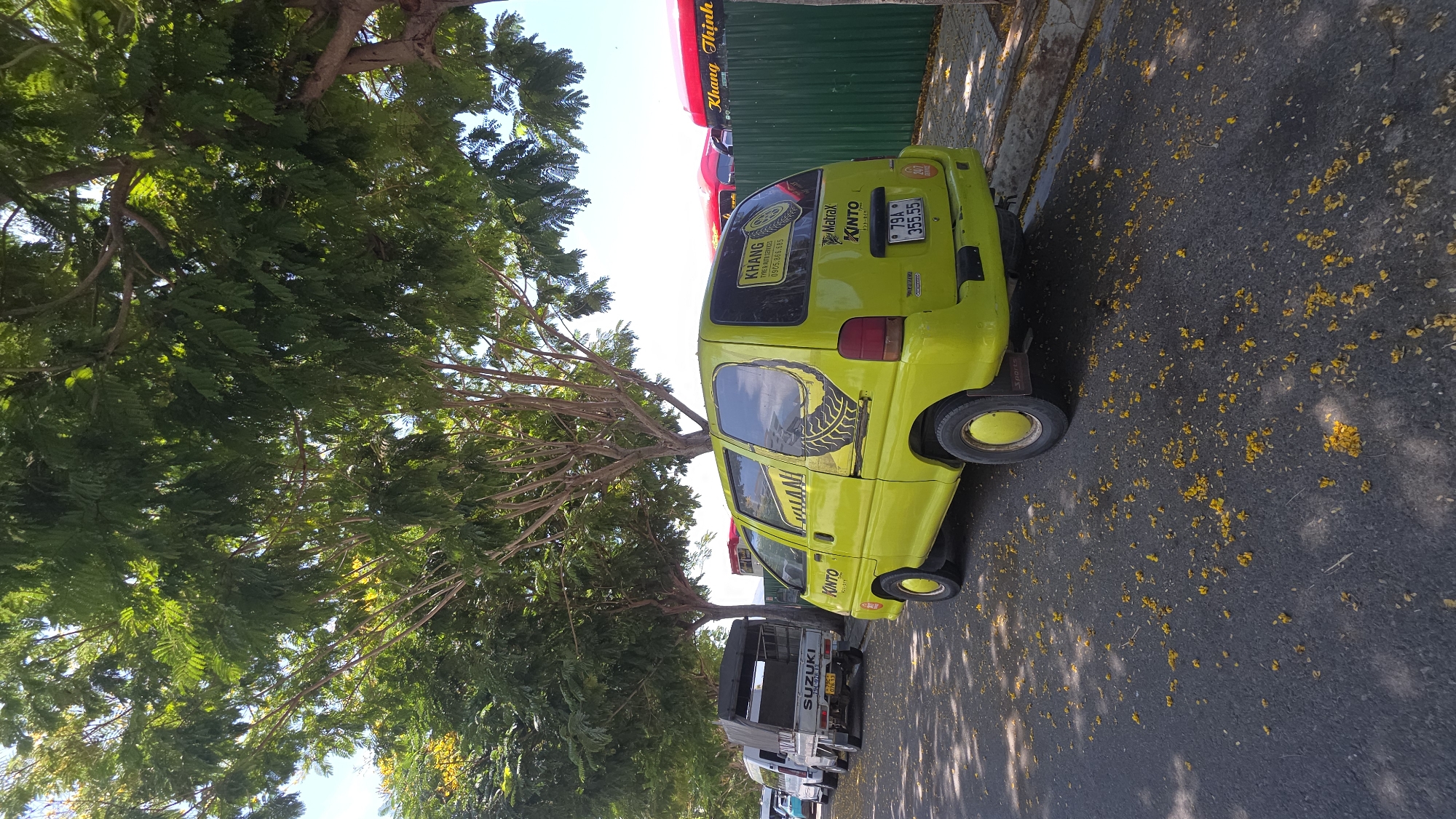 The image captures a street scene in Nam Nha Trang Ward, Vietnam, dominated by a bright yellow Kinto car parked alongside the road. The car prominently displays "KHANG TYRE MUTU SERVICES" and a phone number, suggesting it serves as a mobile advertisement for the business. To the rear of the vehicle, a license plate reads "79A 355.55." Adjacent to the yellow car, several Suzuki trucks are parked in a row, facing the same direction. Overhead, the branches of a large tree spread across the frame, casting shadows on the road. Scattered on the asphalt are numerous small yellow flowers, likely fallen from the tree. In the background, a glimpse of a green corrugated metal wall with red signage reading "Khang Thinh" is visible.