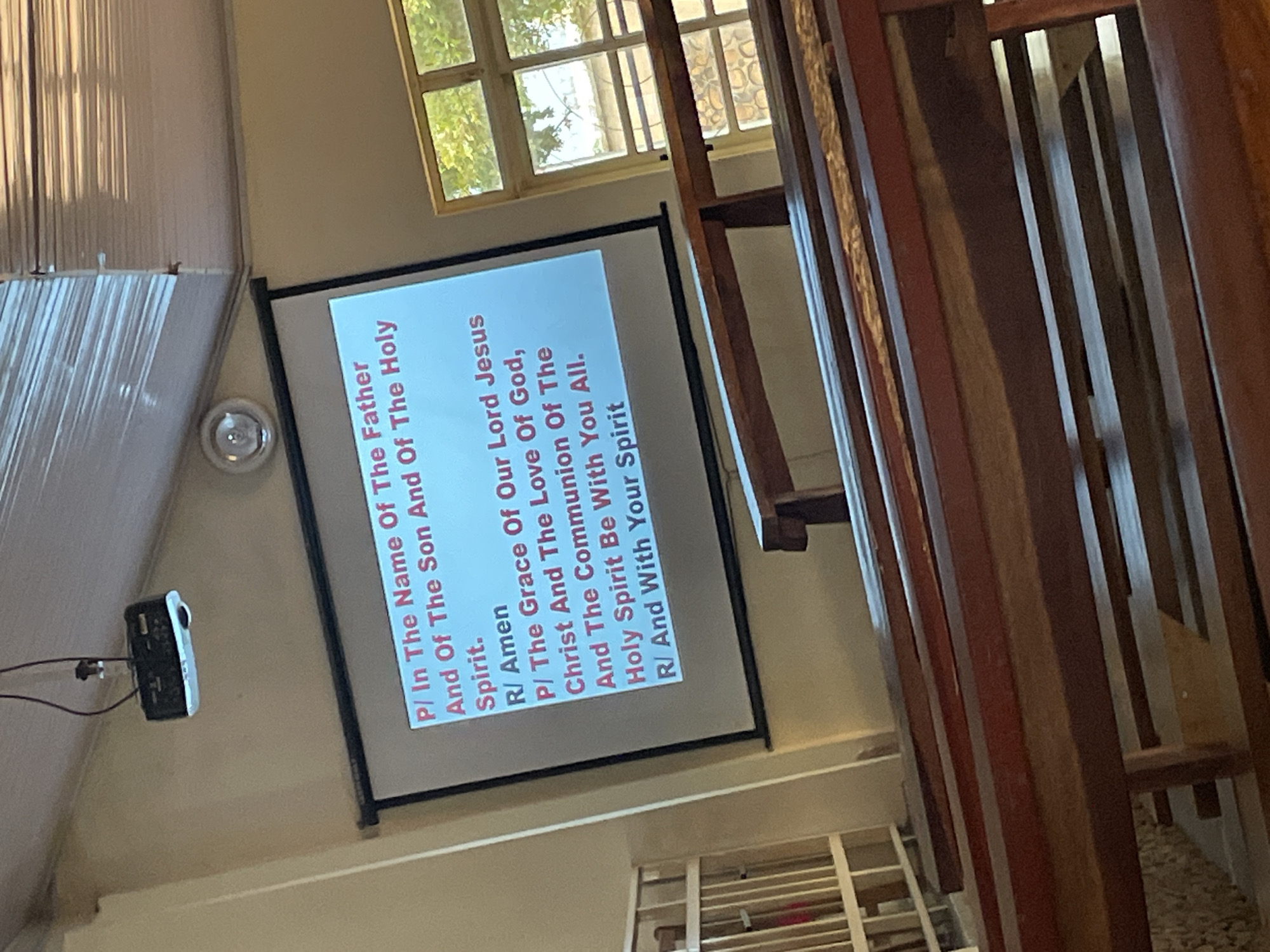 This media file shows a projection screen displaying text that appears to be part of a religious service. The text is written in English and includes phrases like "In The Name Of The Father," "The Grace Of Our Lord Jesus Christ," and "Holy Spirit." The screen is mounted on a wall in a room that appears to be a place of worship, possibly a church, given the religious text.
To the left of the screen, a small black projector is visible, connected by wires. A circular light fixture is also present on the wall near the projector. The room has large windows with blinds on the left side, suggesting it might be daytime. On the right side of the image, a wooden structure, possibly a pulpit or part of the church's architecture, is visible. The overall setting suggests an indoor religious gathering. The text on the screen is clearly legible, and the image is well-lit, though the angle of the shot causes the screen and surrounding elements to appear tilted. No people are visible in the frame. The provided location context of Bar Jirgi Summa, Nigeria, places this scene in a specific geographical setting.