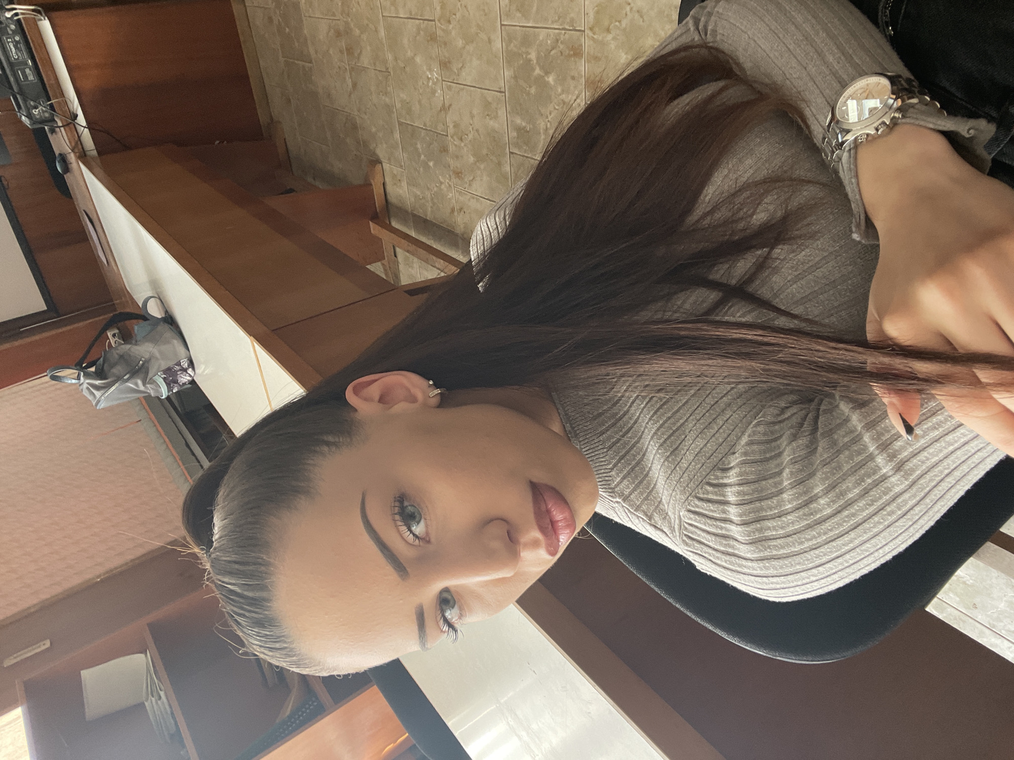 A young woman is shown from a high angle looking up at the camera. Her dark hair is long and flows over her right shoulder. She wears a gray ribbed top and a watch on her left wrist. The background includes a tiled wall, a wooden shelf unit, and part of what appears to be a room interior with cabinets. There is no indication of Veliko Tarnovo, Bulgaria in the image.