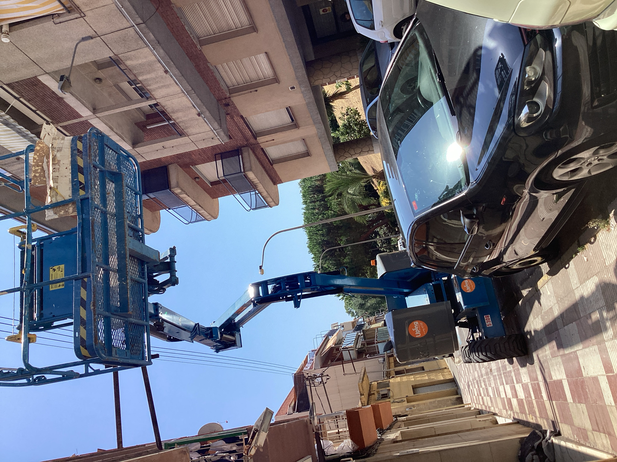 In Blanes, Spain, a blue aerial work platform is parked on a sidewalk, its boom extended towards a building. The platform is positioned next to a black Volkswagen Golf. The sidewalk is paved with red and white square tiles, and shadows from the platform and buildings are cast across it. In the background, a multi-story building with brick and concrete facades stands, featuring balconies and windows with closed blinds. Trees and greenery are visible behind the building, and the sky is clear and blue. The scene appears to be a street in a residential or commercial area, possibly during daytime work or maintenance.