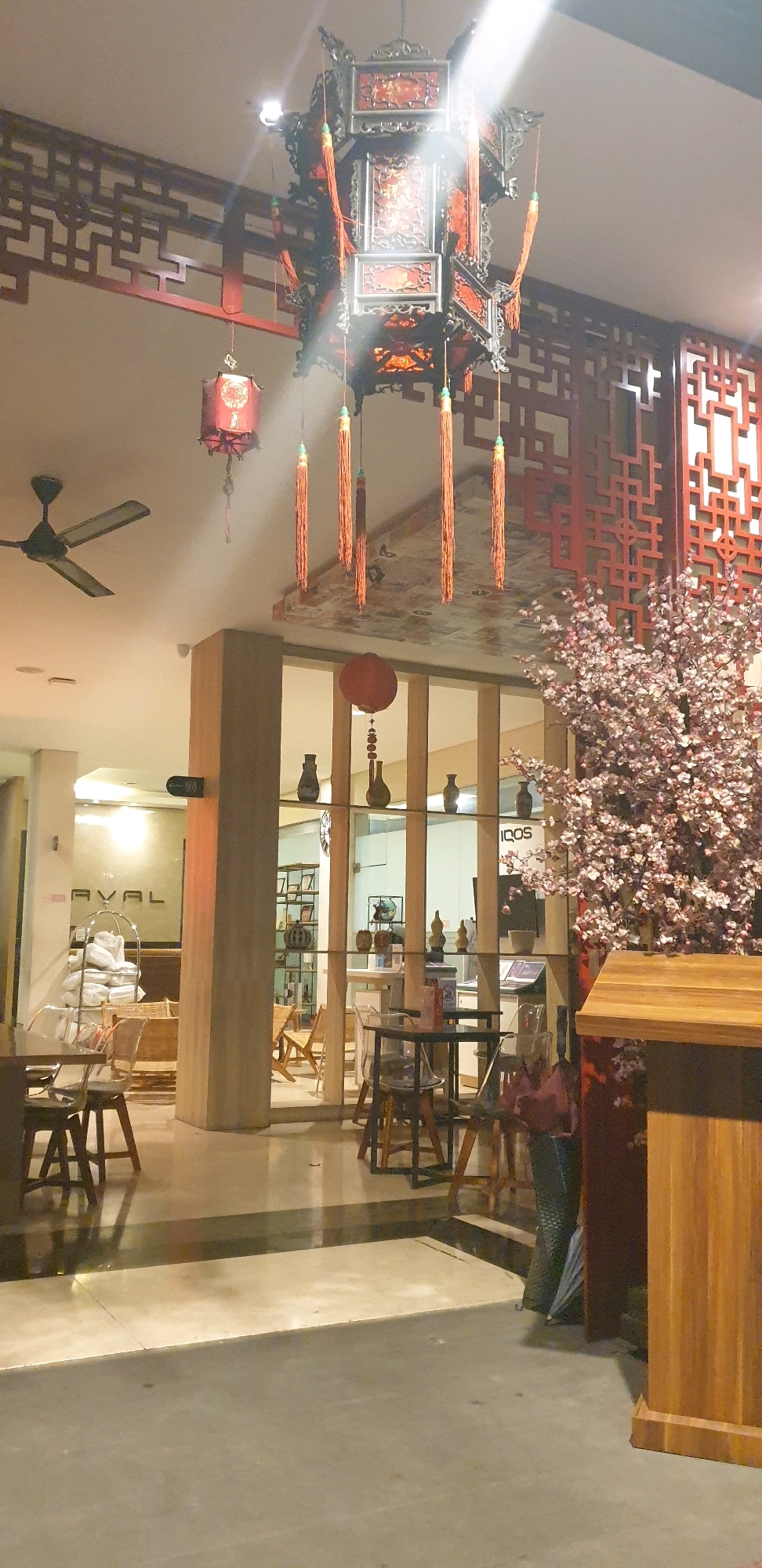 The image shows an interior scene with Asian-inspired decor, likely a restaurant or cafe, with a large, ornate hanging lantern in the center. The lantern is red and black with intricate details and tassels. To the left, a ceiling fan is visible. A smaller red lantern hangs from the ceiling to the left of the main lantern. Red wooden lattice work is present on the wall and ceiling. In the foreground, there are tables and chairs. To the right, a decorative tree with pink blossoms stands next to a wooden podium. Through a glass partition, shelves with vases and other decorative items are visible, along with chairs and tables inside. The flooring is a polished concrete or stone. The overall lighting is warm and ambient.