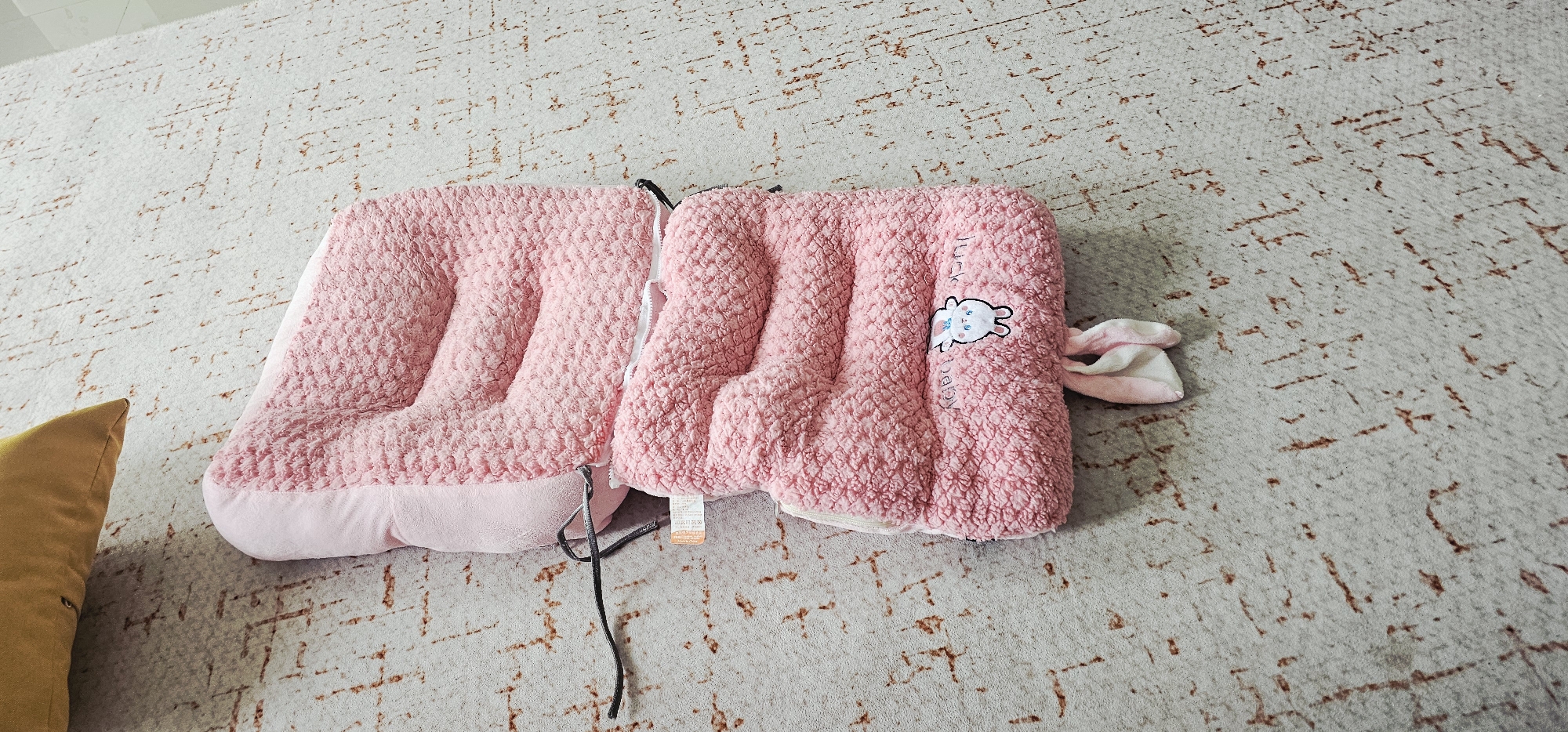 A pink, plush pet bed is positioned on a textured, cream-colored rug with subtle brown markings. The bed appears to be folded in half, revealing a white zipper closure. One side of the bed features a cartoon bunny illustration and what appears to be text, possibly "lucky baby" written vertically. Attached to the bed are two small, pink and white bunny ears. A dark cord extends from the underside of the bed. To the left of the pet bed, a corner of a mustard yellow pillow is visible. The setting is indoors, on a rug, likely in a home or pet-related establishment. There is no visible indication of the location being in Chongqing High-tech Zone, China.
