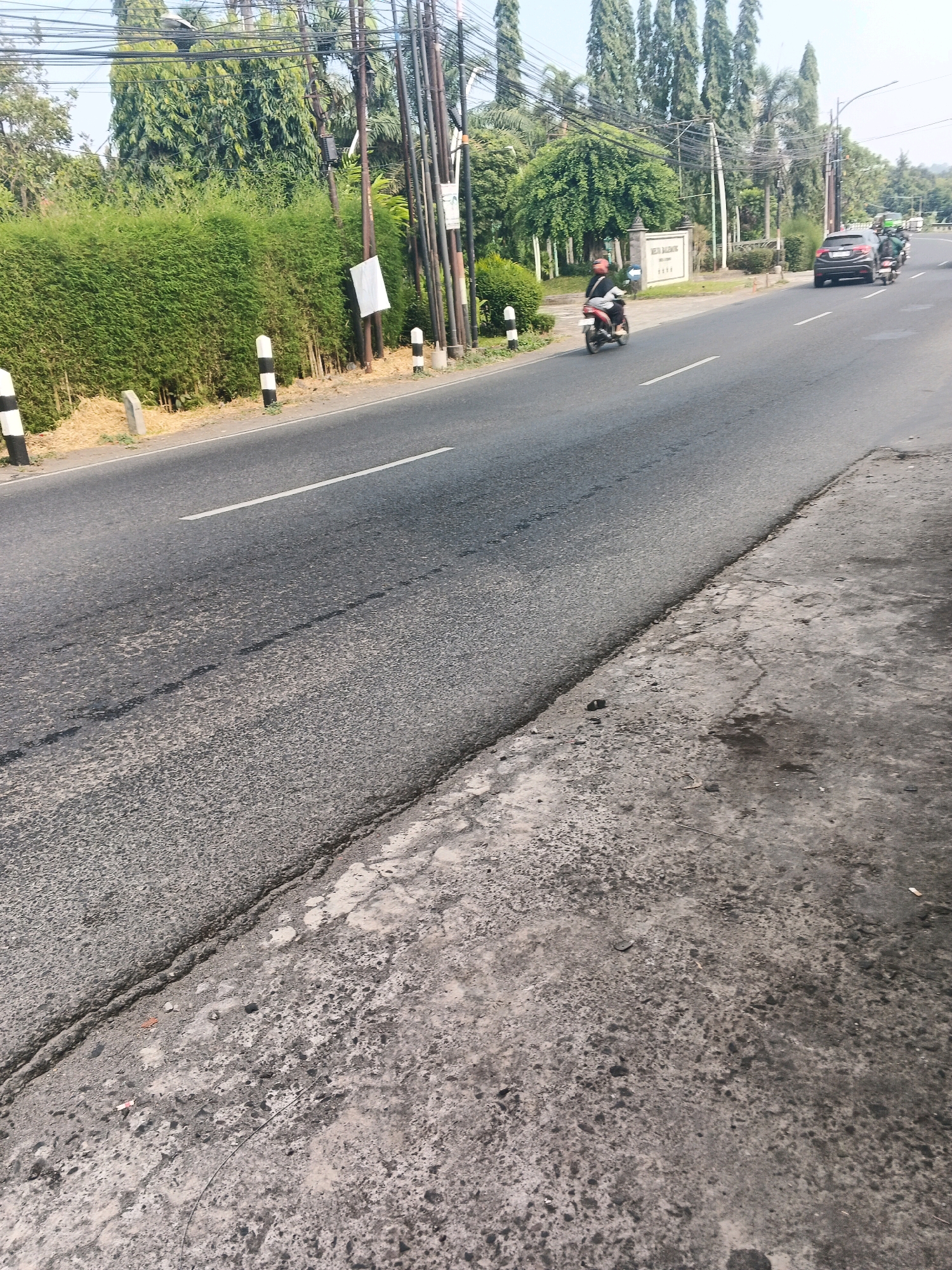The image shows a road with a white line marking a lane, and a grey asphalt surface with some cracks and debris. On the left side of the road, there is a thick green hedge and some black and white bollards. Further down the road, there is a sign with text that appears to be in Indonesian, possibly indicating a location within Ungaran, Indonesia. A person on a scooter is riding in the middle lane. In the background, there are trees and utility poles with wires. On the right side of the road, a dark-colored car is parked or driving. The overall scene depicts a street environment in a tropical or subtropical region, likely during daylight hours given the lighting conditions.