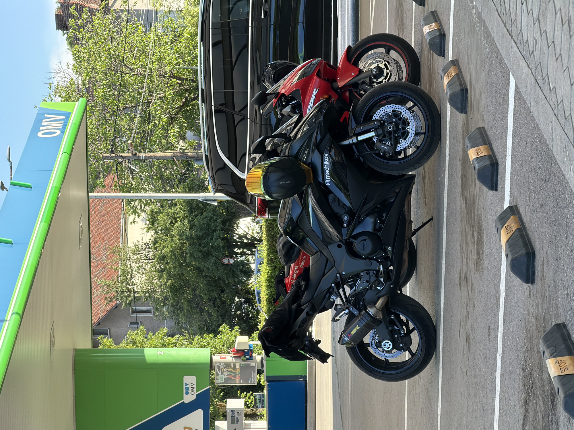 The image shows two motorcycles parked in front of a green and blue OMV gas station. One motorcycle is black and the other is red. A black van is parked behind the motorcycles. The ground is paved with parking space indicators. The picture was taken during daytime in Sofia, Bulgaria.