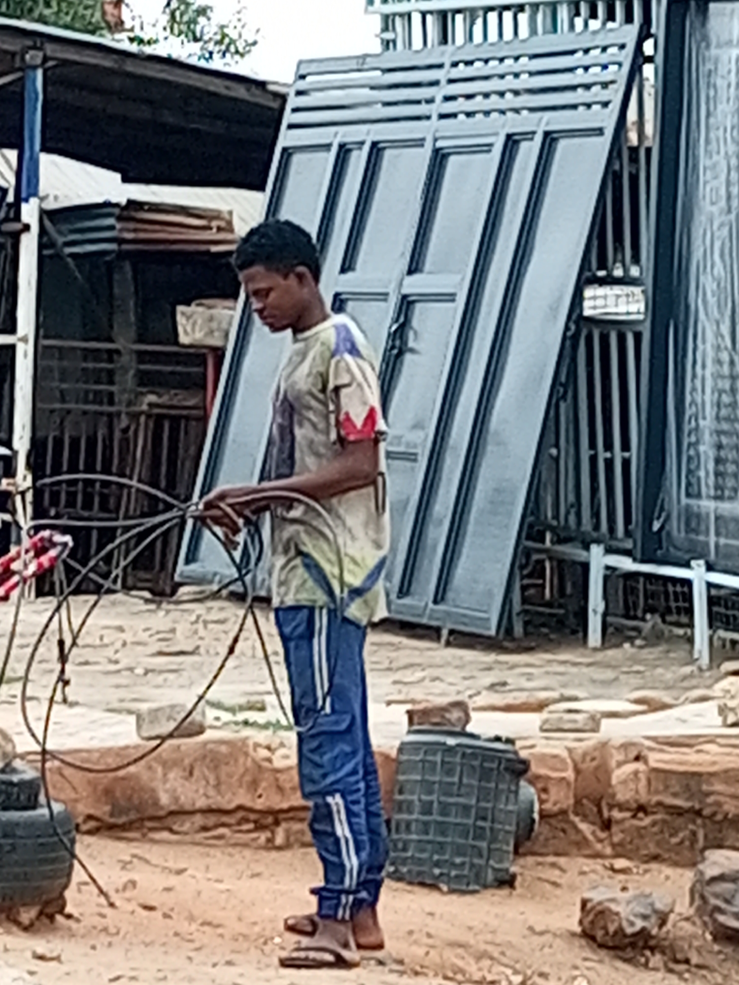 In Garko, Nigeria, a young person, appearing to be a male, stands in a sandy, outdoor area. He is wearing a light-colored t-shirt with red and blue accents on the sleeves and collar, and blue pants with white stripes down the legs. His feet are clad in brown sandals. The person is holding a bundle of black wires or cables in both hands. Behind him, a large, gray, metallic structure, possibly a gate or large panel, leans against a background of more metal structures and corrugated metal sheeting. The ground is a mixture of sand and dirt, with scattered rocks and some larger, rough-hewn blocks. To the left of the person, a black tire is partially visible, and further back, there is a dark metal fence and a structure with a dark roof. To the right, a dark, caged object sits on the ground. The overall scene suggests an industrial or workshop environment.