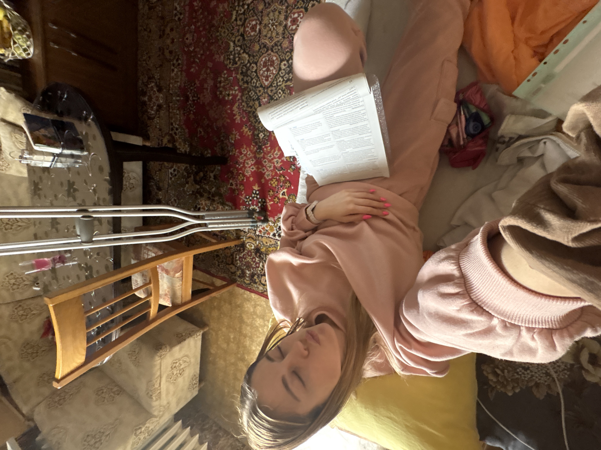 A young woman is lying on a bed covered with various pillows and blankets. She is wearing pink sweatpants and a matching sweatshirt. She is holding a document in her hands. A pair of crutches is visible next to a small wooden table with various items on it. The bed is on a carpet in a room with floral-patterned furniture. The image was likely taken indoors. There is no information in the image indicating the location is in Sofia, Bulgaria.