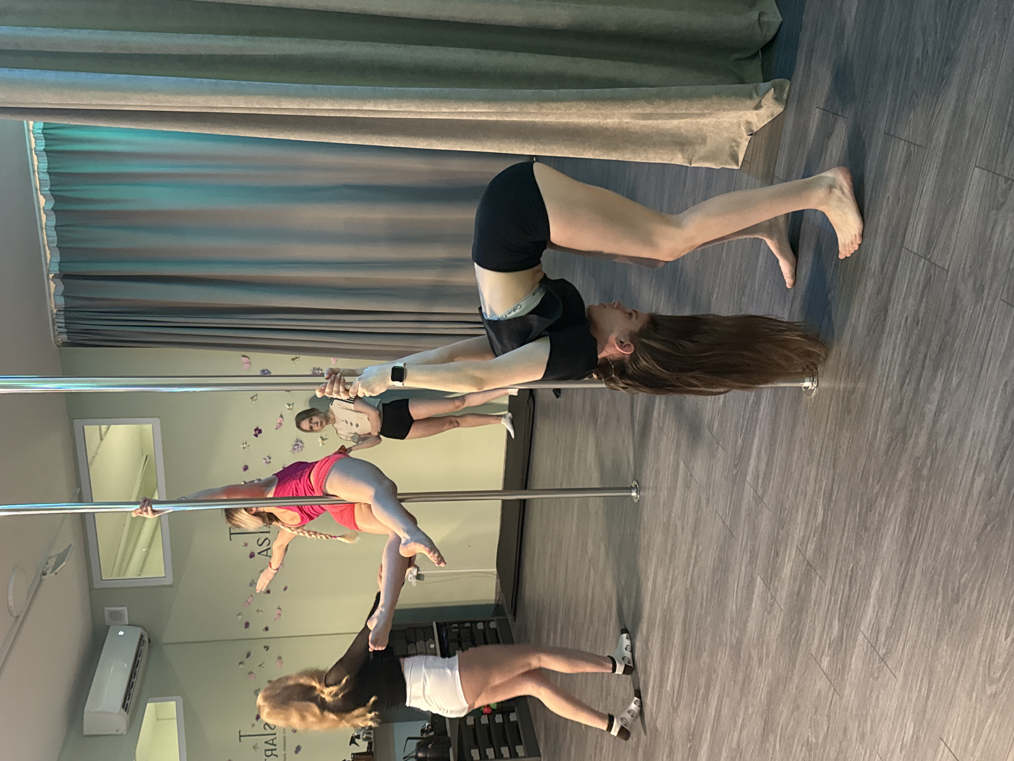 Captured at a pole dancing studio in Batumi, Georgia, the photograph features four women engaged in various pole exercises. The studio is a light-filled space with gray wood-look flooring, green-toned curtains, and walls adorned with butterfly decals and the words "STAR TASI." Three silver-colored poles are visible, each occupied by a woman in a different pose. In the foreground, a woman is positioned upside down on the pole, her legs extended, supporting her body with her hands on the pole. She wears black shorts, a black top, and a fitness tracker on her wrist. To the left, a woman in a pink top and shorts is performing a static pose, with both hands gripping the pole. A third woman is seen in the background, near the middle pole, wearing a light-colored top and dark shorts. In the lower left corner of the image, a fourth woman is standing with one leg straight and one bent, wearing white shorts and a black top. The room contains other details, including a mirror, an air conditioning unit, and some fitness equipment in the background.