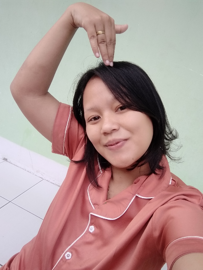 A smiling young woman poses in a casual setting, likely indoors. She is wearing a reddish-orange pajama-style top with white trim. Her hair is short, dark, and styled with bangs. Her right arm is raised above her head, with her fingers nearly touching her hair, revealing a gold ring on her finger. The background is a light green wall, with a hint of white tiles at the bottom.