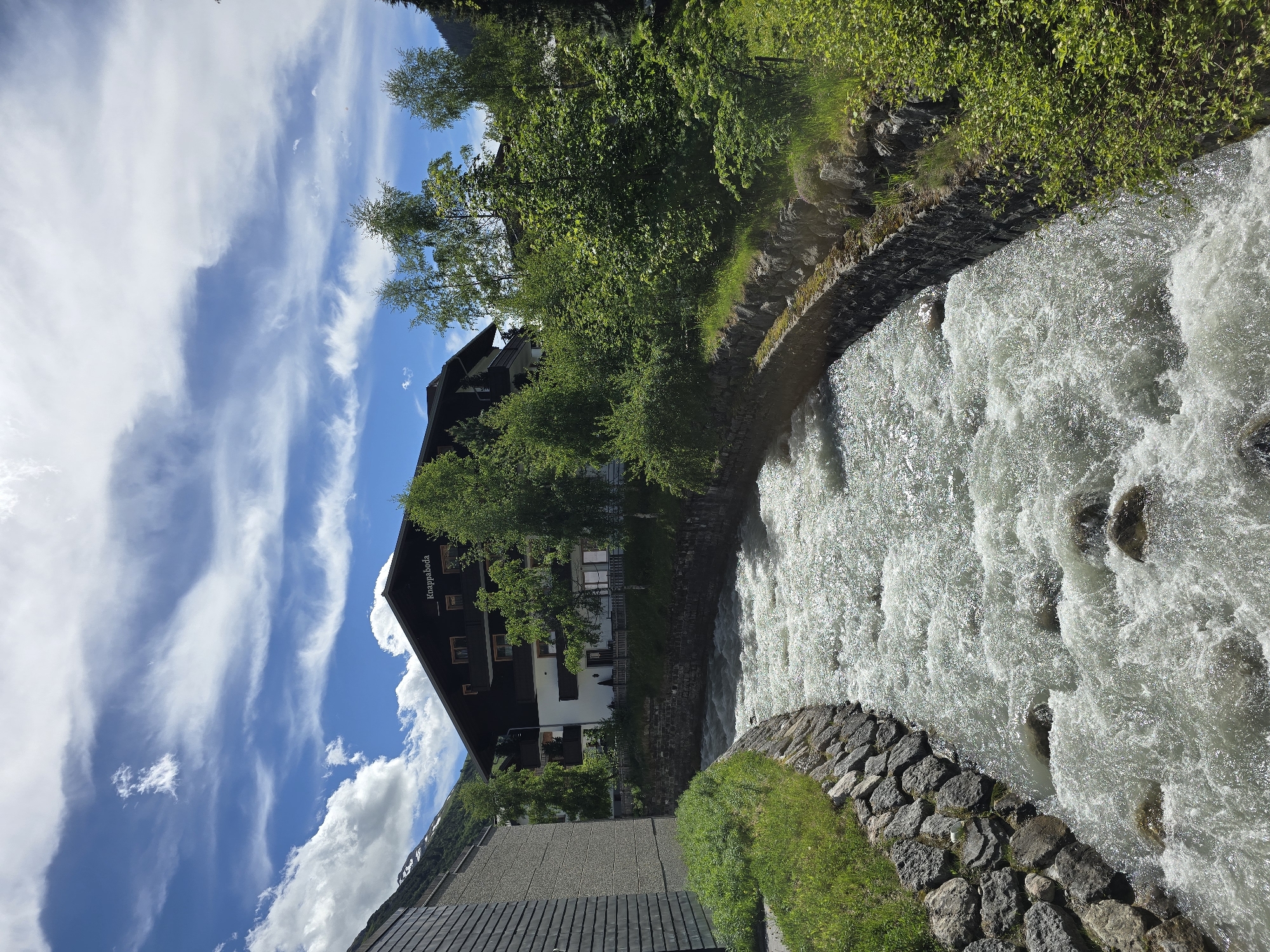 The image shows a fast-flowing river with white water rapids, bordered by stone walls on both sides. On the right bank, lush green foliage covers the embankment. On the left bank, there is a grassy area and the side of a modern building. Across the river, a dark wooden building with multiple balconies and windows is visible, bearing the sign "Knappaboda". Behind this building, more trees and vegetation are present, with a mountain slope in the distance. The sky above is blue with scattered white and wispy clouds. The scene is likely captured in Lech, Austria, given the architectural style and mountainous surroundings typical of the region.
