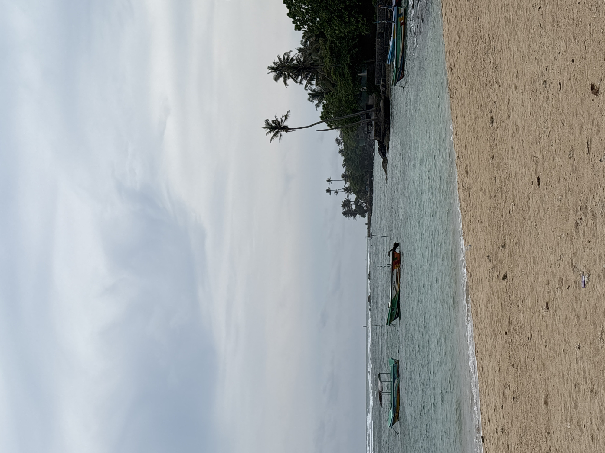 The image depicts a beach scene in Weliweriya, Sri Lanka. The sky is overcast with light grey clouds. Along the shoreline, there is a sandy beach on the right side. To the left of the beach, the water is a light blue-green. Palm trees and lush green vegetation line the edge of the beach. Several small, narrow fishing boats are in the water, close to the shore. One boat has a person on board.