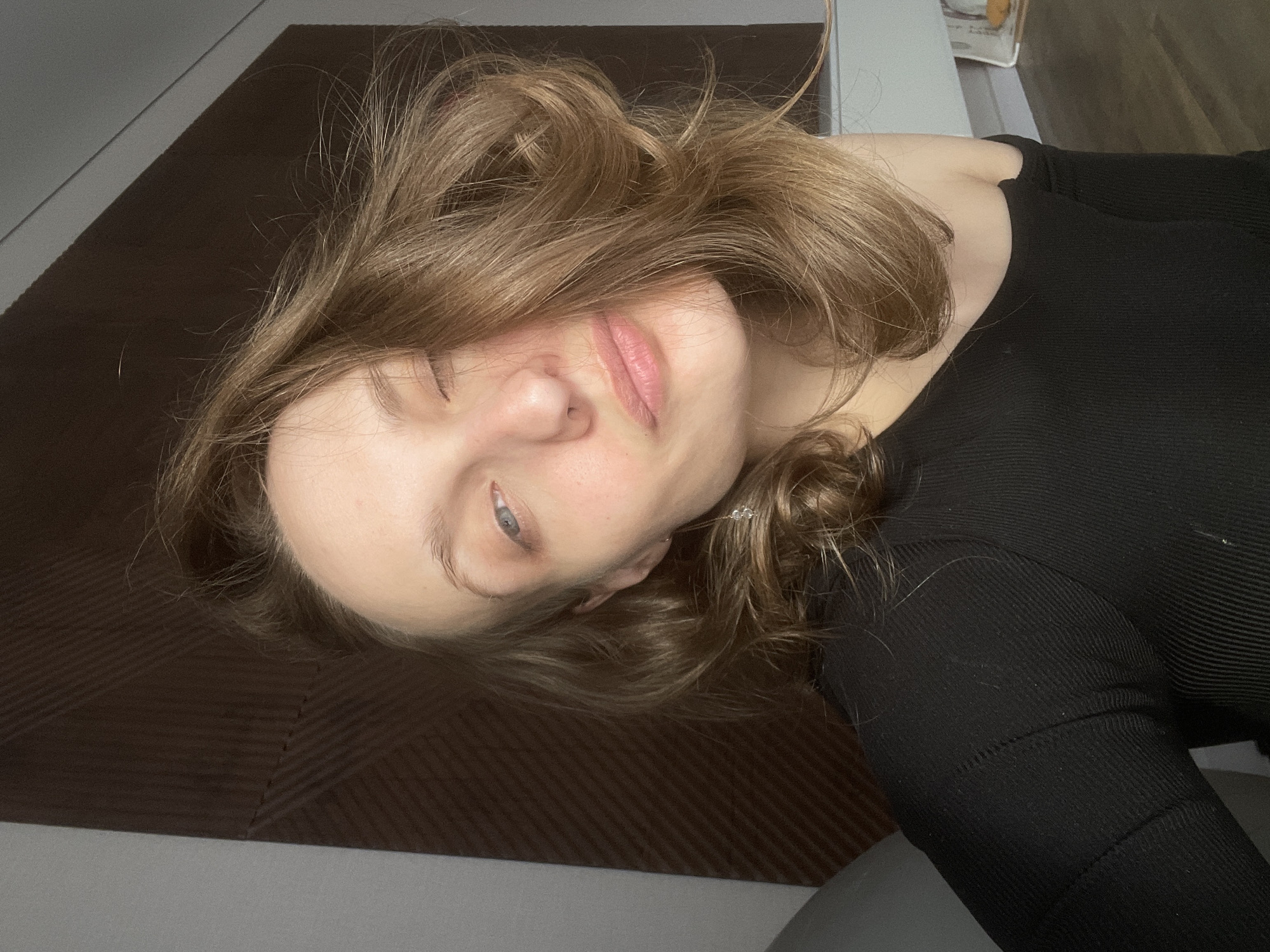 A close-up, tilted portrait features a young woman lying down indoors. Her light brown, wavy hair, styled with some volume, frames her face, with strands falling over her forehead and cheeks. She looks towards the camera with a soft, relaxed expression; one eye is partially open and gazing forward, while the other appears almost closed. Her lips are slightly parted, showing a light pink lip color.
She is wearing a black, ribbed long-sleeved top or dress, visible on her shoulder and arm. A small, subtle ear cuff or earring with clear beads adorns her right ear.
The background primarily consists of dark brown, interlocking floor mats, which appear to be textured foam. A light gray surface, possibly a wall or another floor section, is visible in the upper left and lower right portions of the frame. The lighting is soft and even, suggesting an indoor setting during the day or under artificial illumination. No specific textual information or unique architectural cues within the image confirm the location, though the context provided places it in Подшивалово, Russia.