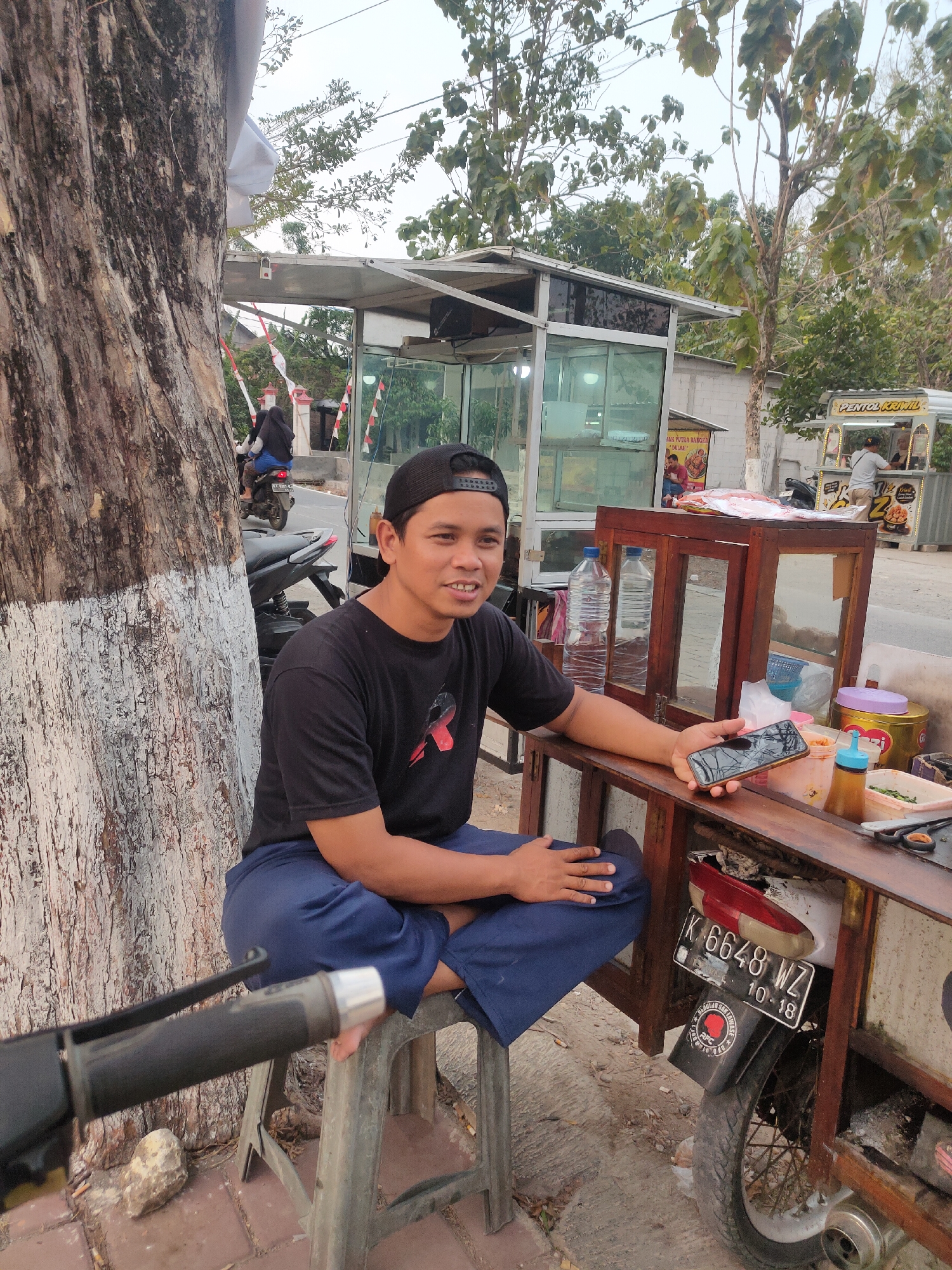 A man wearing a black t-shirt and blue pants is sitting on a stool next to a wooden cart. He is holding a mobile phone in his right hand and looking at the camera. The cart is filled with various items, including bottles of water, containers, and utensils. In the background, there is a food stall with a glass display case, and people are seen ordering food. A motorcycle is parked behind the cart. The scene appears to be a street in Karanglangu, Indonesia, as indicated by the license plate on the motorcycle.