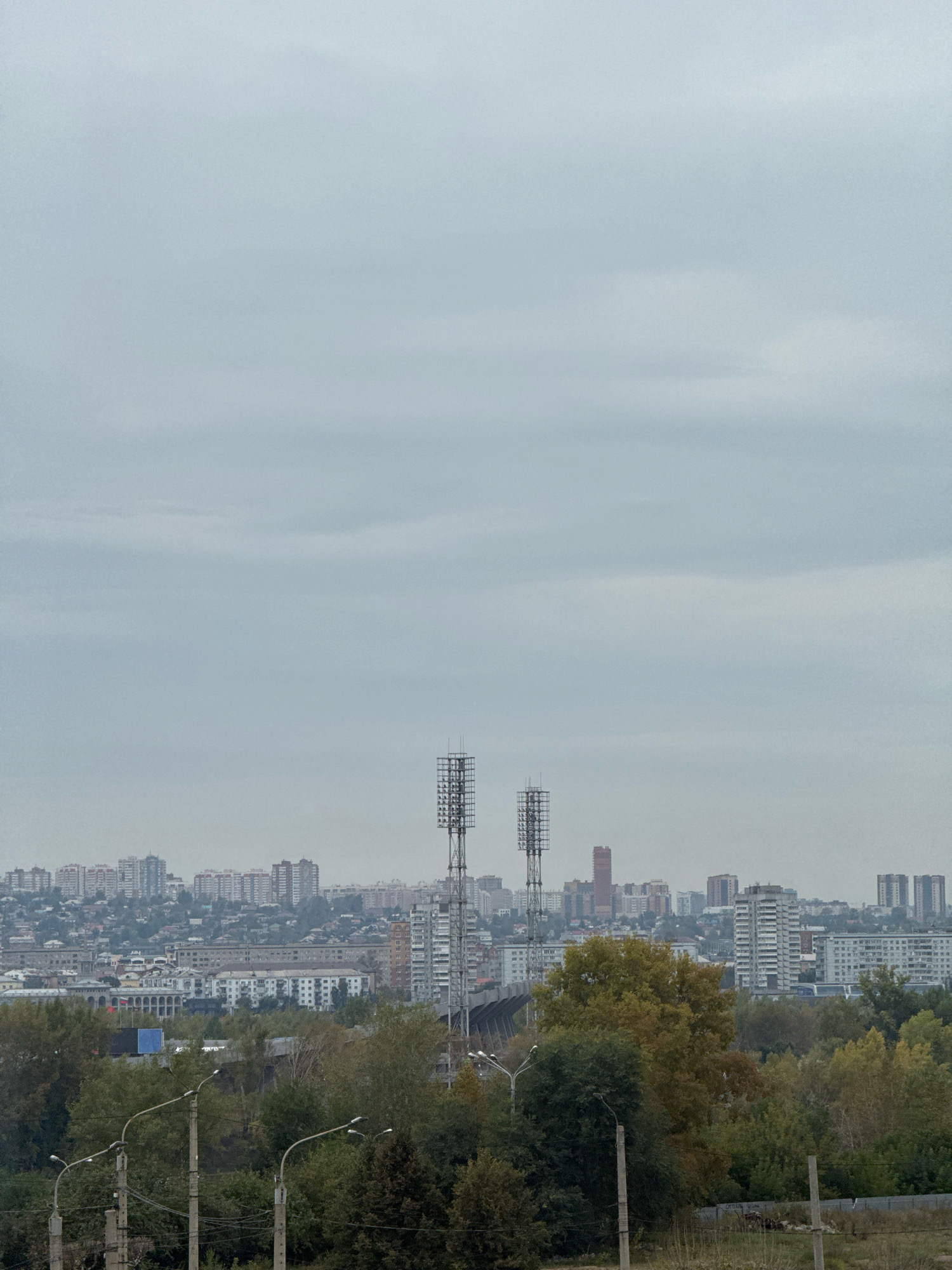 The image shows a panoramic view of a city on a cloudy day, likely in Krasnoyarsk, Russia, as indicated by the architecture and infrastructure. In the foreground, several concrete lampposts are visible, some with multiple lights, amidst dense green foliage and trees, some of which are exhibiting autumn colors of yellow and orange. Beyond the trees, a cityscape unfolds with a variety of buildings, including multi-story apartment blocks and smaller residential structures. Two prominent communication towers, appearing to be lattice towers, stand tall in the middle ground. One of these towers is closer and appears to be part of a stadium or sports complex structure, with supporting beams visible. In the background, the city extends towards the horizon, with a mix of modern high-rises and older, lower buildings. The sky is overcast with heavy, gray clouds, obscuring any direct sunlight and contributing to a muted color palette. There are no visible people or animals in the frame. The overall impression is an urban environment with elements of nature, viewed from a slightly elevated position.