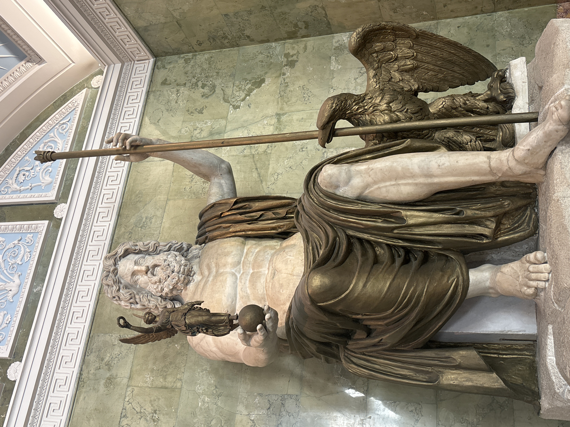 The image depicts a sculpture of Zeus, the king of the gods in Greek mythology. He is shown lying down, with his body angled diagonally across the frame. His muscular torso is visible, as well as his legs and feet. He is draped in a golden cloth that emphasizes his powerful physique. In his right hand, he holds a small, round object, possibly a globe or an apple. His left hand grasps a long, golden staff that extends upwards and to the left, where it is held by an eagle perched on his shoulder. The eagle is rendered in great detail, with its wings spread and its beak open. Zeus has a full beard and flowing hair, characteristic of his depiction in art. The sculpture is made of what appears to be marble and bronze, with the marble forming the main body of Zeus and the bronze used for the drapery, the eagle, and some of his accessories.
The setting appears to be an indoor space, possibly a museum or a grand hall, with green, marble-like walls and ornate white architectural details along the top of the wall and ceiling. The architectural elements include a Greek key pattern and decorative molding. The lighting is even, suggesting an artificial light source. The overall impression is one of grandeur and classical artistry. The sculpture likely represents Zeus in a moment of repose or contemplation, perhaps after a significant event. There is no visible text in the image. The orientation of the image is vertical, as if the viewer is looking up at the sculpture from a lower vantage point.