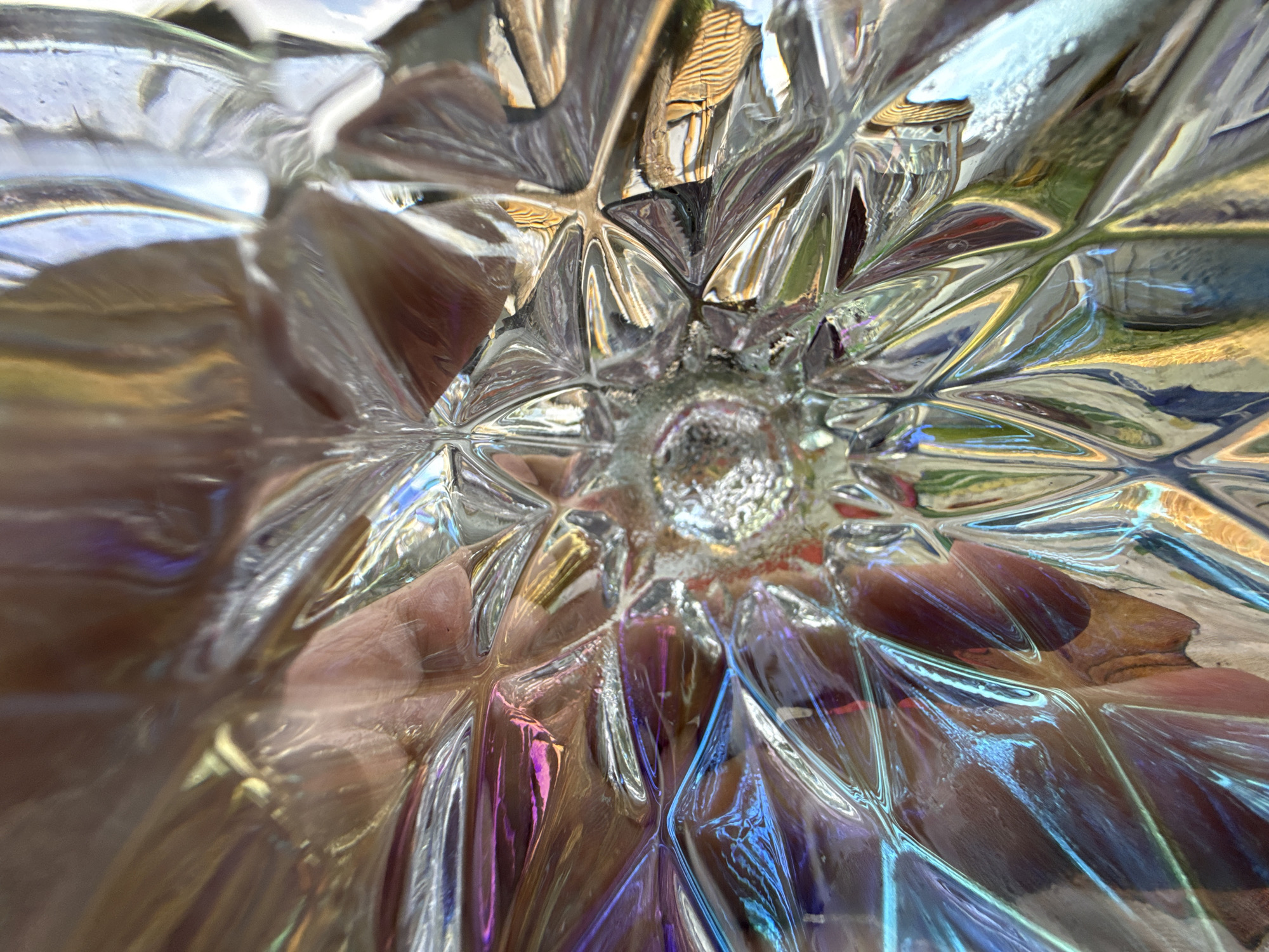 The image shows a close-up view of a textured glass object, possibly a vase or a decorative piece, with a hand visible through its transparent surface. The glass has intricate facets that refract light, creating a spectrum of colors, including blues, purples, and yellows. The texture of the glass creates a starburst pattern radiating from the center. The hand appears to be gently pressing against the inside of the glass. The background is out of focus but suggests an indoor environment with reflected light and possibly other objects. The scene is set in Sevan, Armenia.