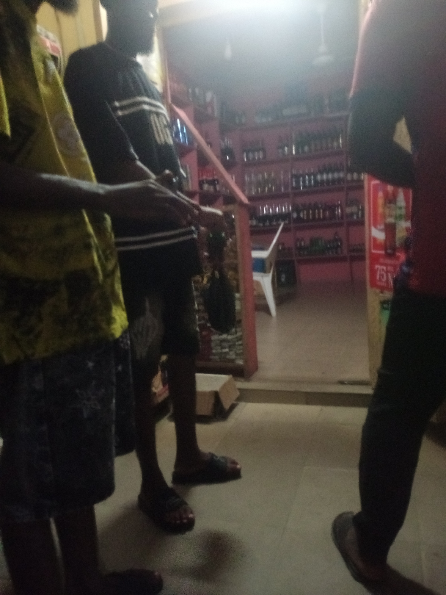The image shows three individuals standing in what appears to be a convenience store or a small shop in Jalingo, Nigeria. The store is stocked with various items on shelves, including bottles, suggesting it might sell beverages or other packaged goods. One individual on the left is wearing a yellow shirt with a patterned design and dark shorts. In the center, another person is wearing a black long-sleeved shirt with white stripes and dark pants. The third individual on the right is largely obscured, wearing a red shirt and dark pants. The lighting in the store is dim, with some illumination coming from overhead. The floor appears to be tiled. The scene captures a moment of everyday life in Jalingo, possibly a casual gathering or interaction within the shop.