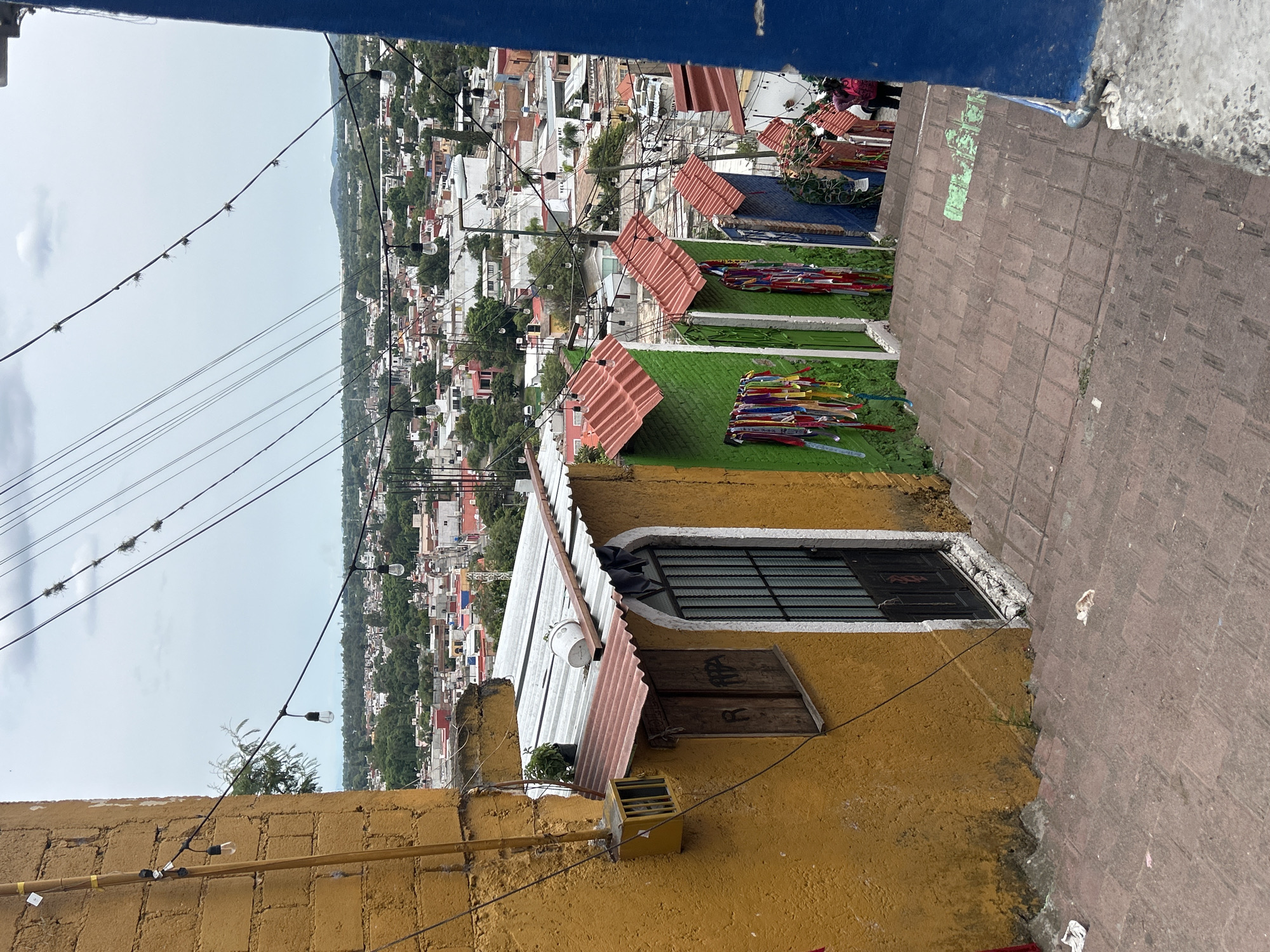 This is a vertical, eye-level shot looking down a set of tiled steps in Atlixco, Mexico. The steps descend past a series of brightly colored, narrow structures, likely small shops or stalls, with corrugated metal roofs. The structures are painted in green, blue, and red. They are decorated with strings of colorful ribbons and what appear to be small flags or banners. The overall scene has a rustic and vibrant feel. On the left, a mustard-yellow building with a rough-textured wall is visible, featuring a window with wooden shutters and a metal-grated door. String lights with exposed bulbs are strung across the scene, casting a soft glow. In the background, a sprawling cityscape with a mix of white, red, and pastel-colored buildings unfolds under a slightly overcast sky. Trees and greenery are interspersed throughout the urban landscape. A few people are visible in the distance, walking down the steps. The foreground is dominated by the red-tiled steps, some of which have a green painted design. There is a small amount of litter on the steps.